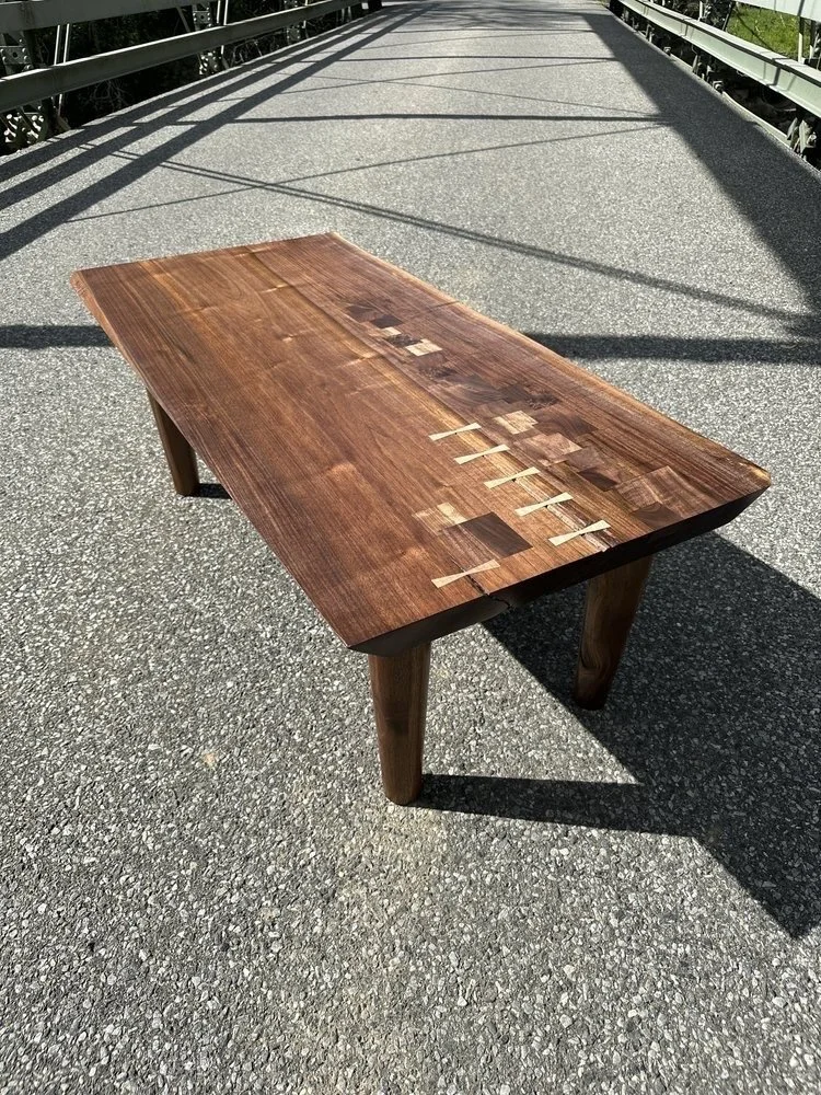 A wooden table placed on a bridge sidewalk with metal railings on either side, casting shadows on the table and the pavement.
