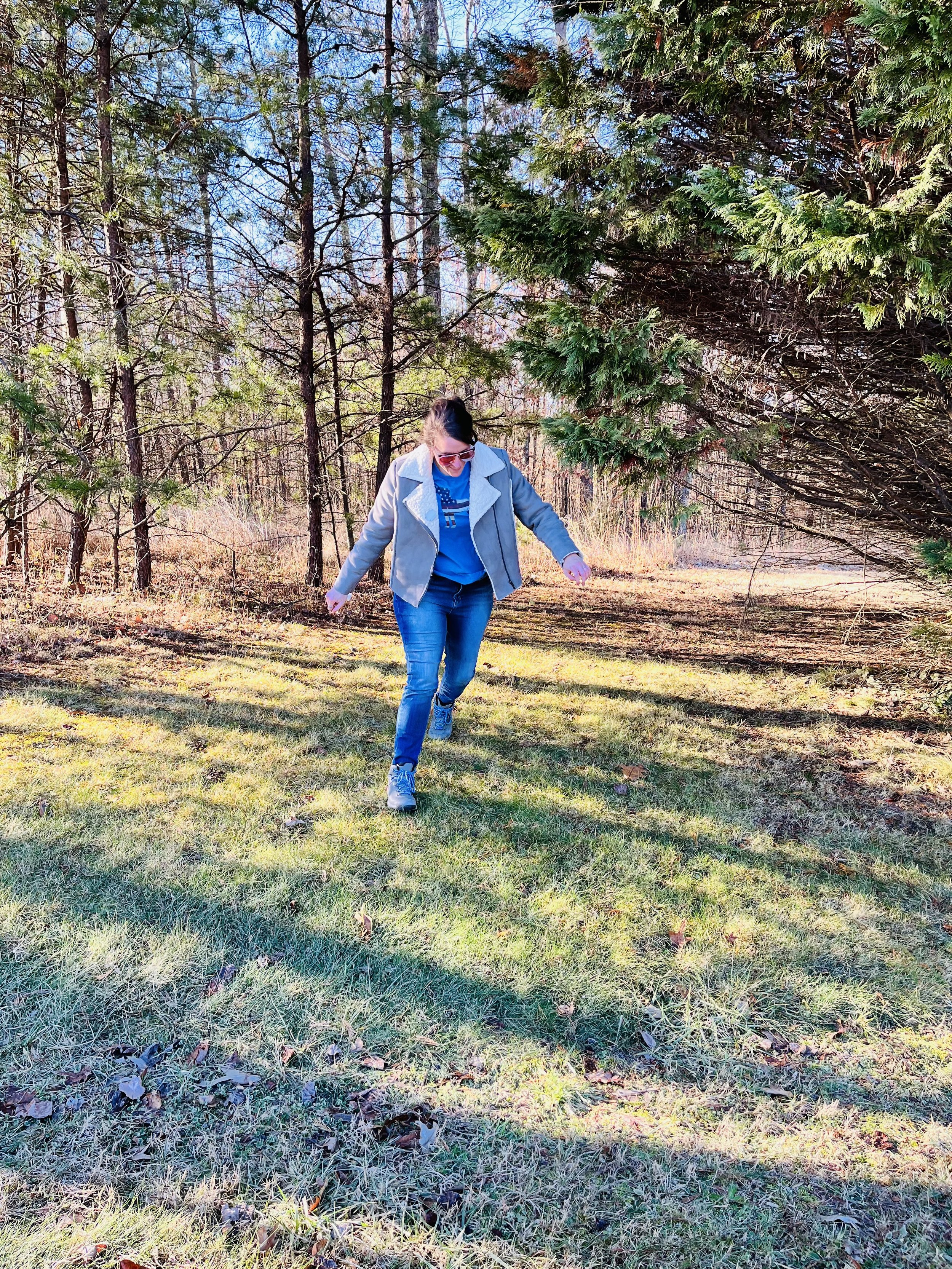A person walking on grass in a forested area on a sunny day, wearing a gray jacket and blue jeans.