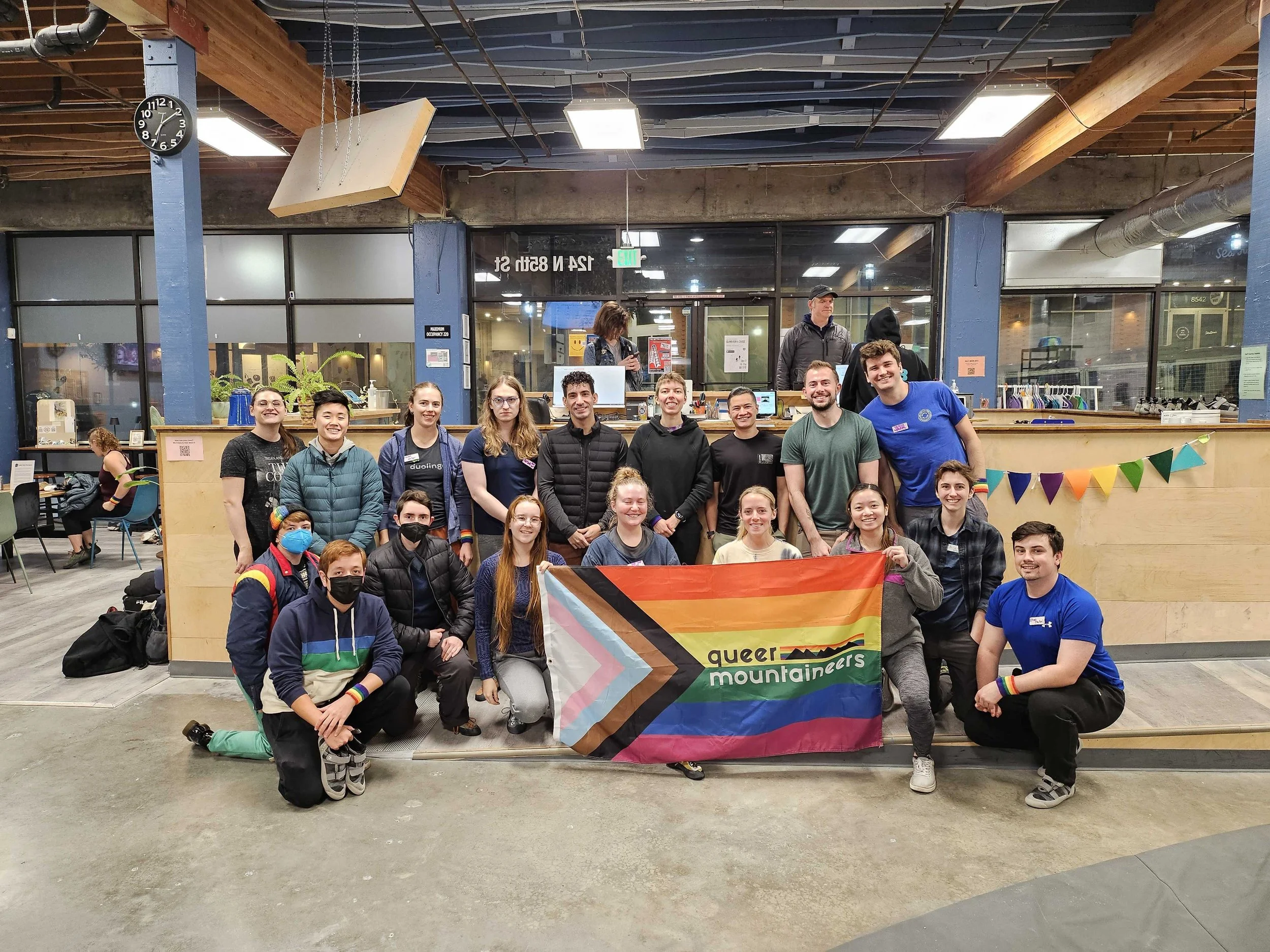 A group of people indoors posing with a "Queer Mountaineers" rainbow Pride flag featuring rainbow and trans pride colors. The setting is a climbing gym with tables and decorations in the background.
