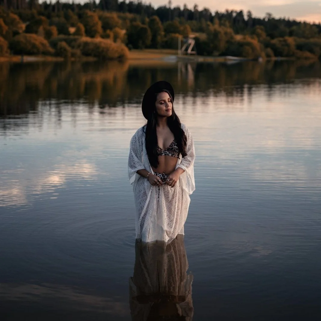 Frau steht im einem Bikini, Badekleid und schwarzem Hut gekleidet bis zu den Waden in einem See inmitten von Natur