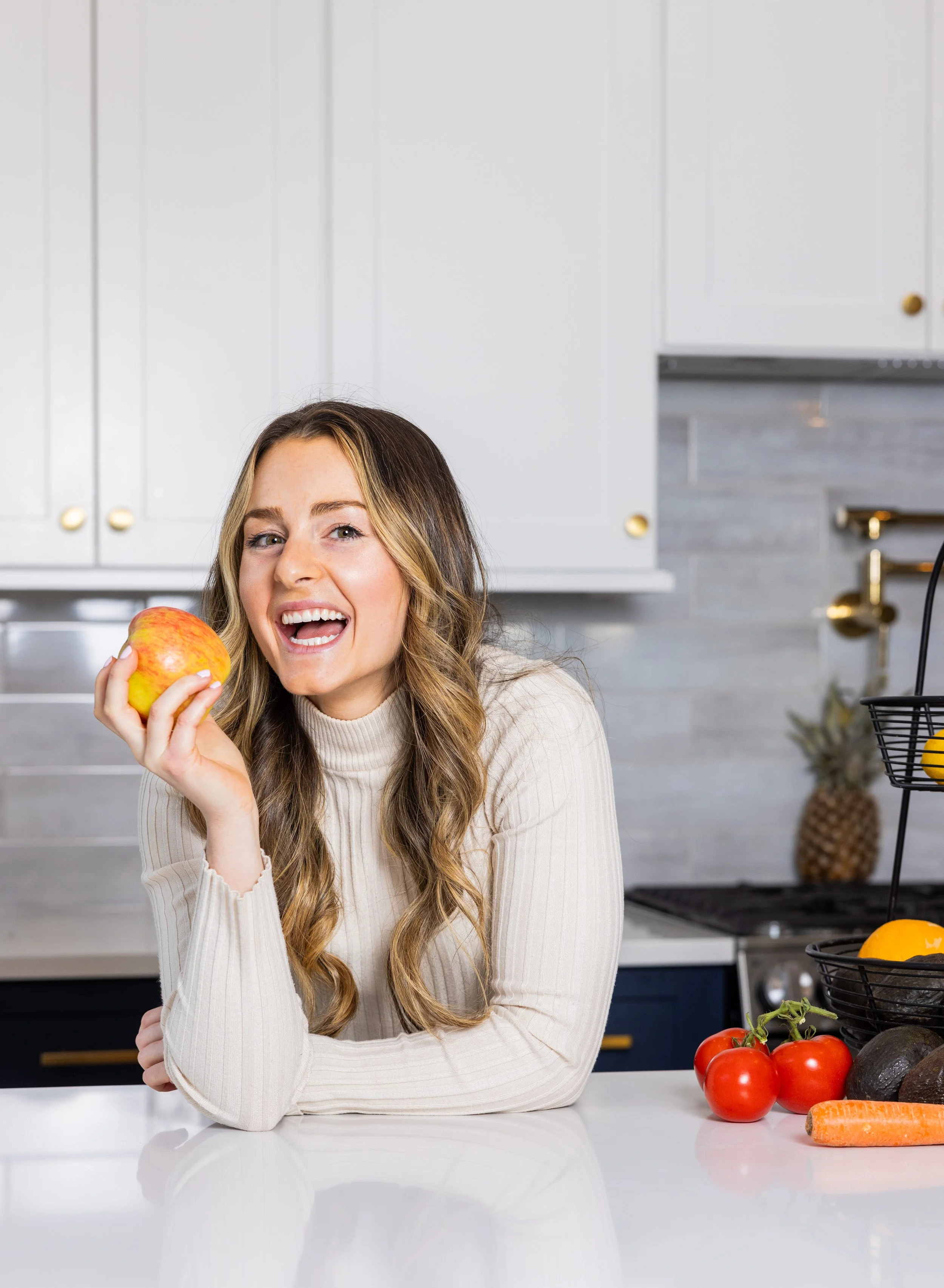 A close-up of Molly Pelletier, RD, smiling and holding a red apple near her face, resting on a kitchen counter with fresh produce in a bright kitchen setting.