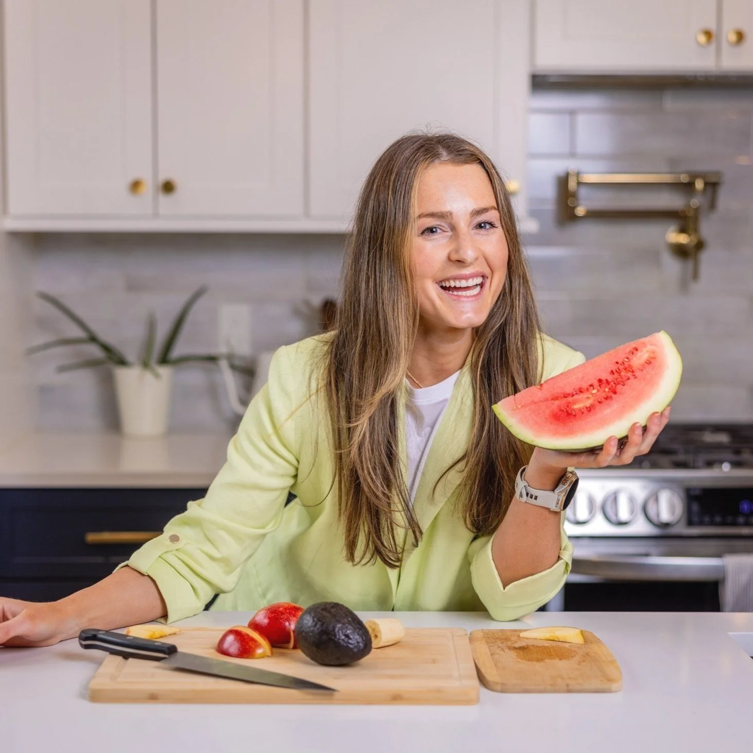 A medium shot of Molly Pelletier, RD, smiling while holding a large slice of watermelon over a kitchen counter with cut fruit in a bright kitchen setting.