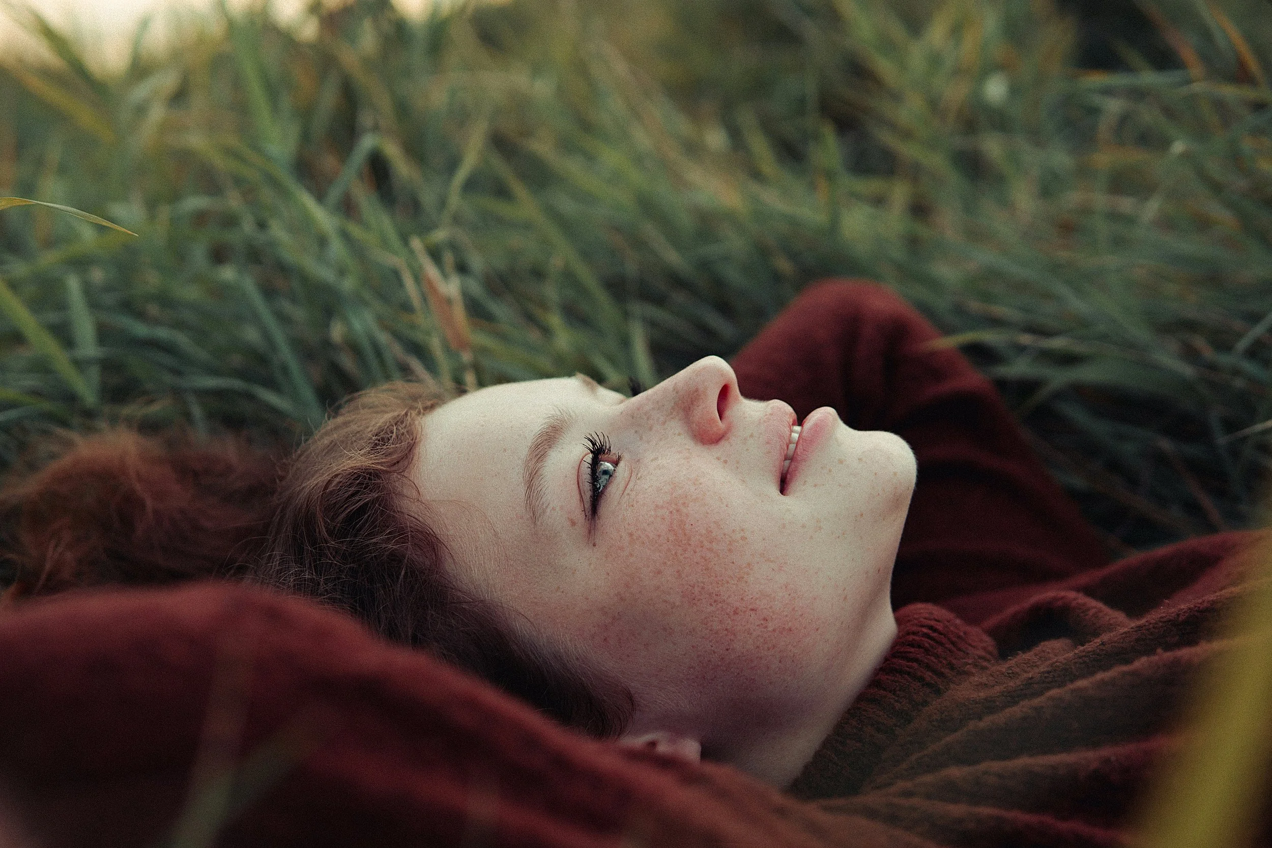 A young woman with pale skin and red hair lying on the ground among green grass, smiling and looking upward.
