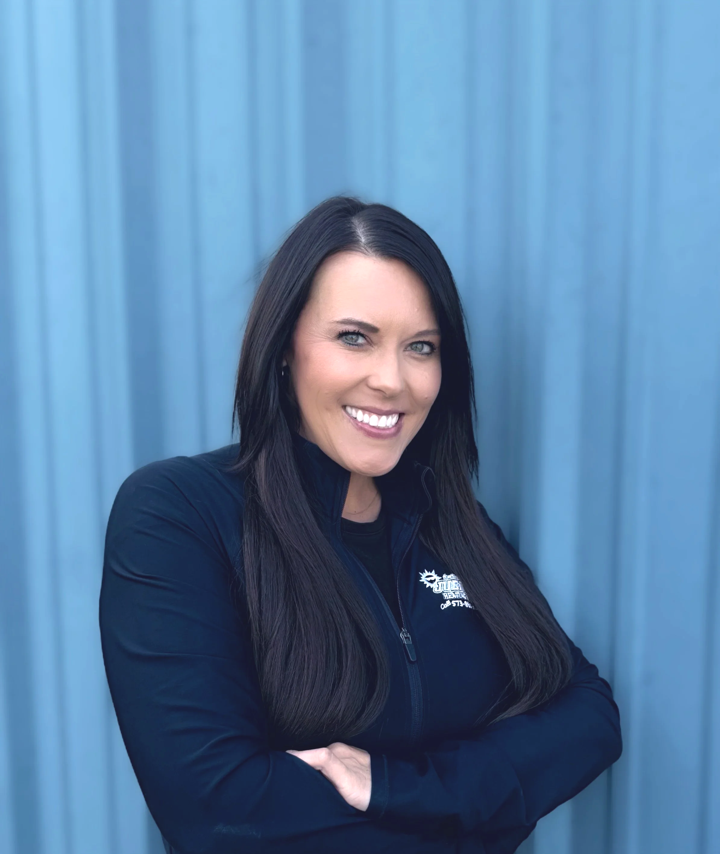 A woman with dark brown hair, blue eyes, and wearing a blue jacket and gray shirt, standing in front of a blue metal wall, smiling at the camera.
