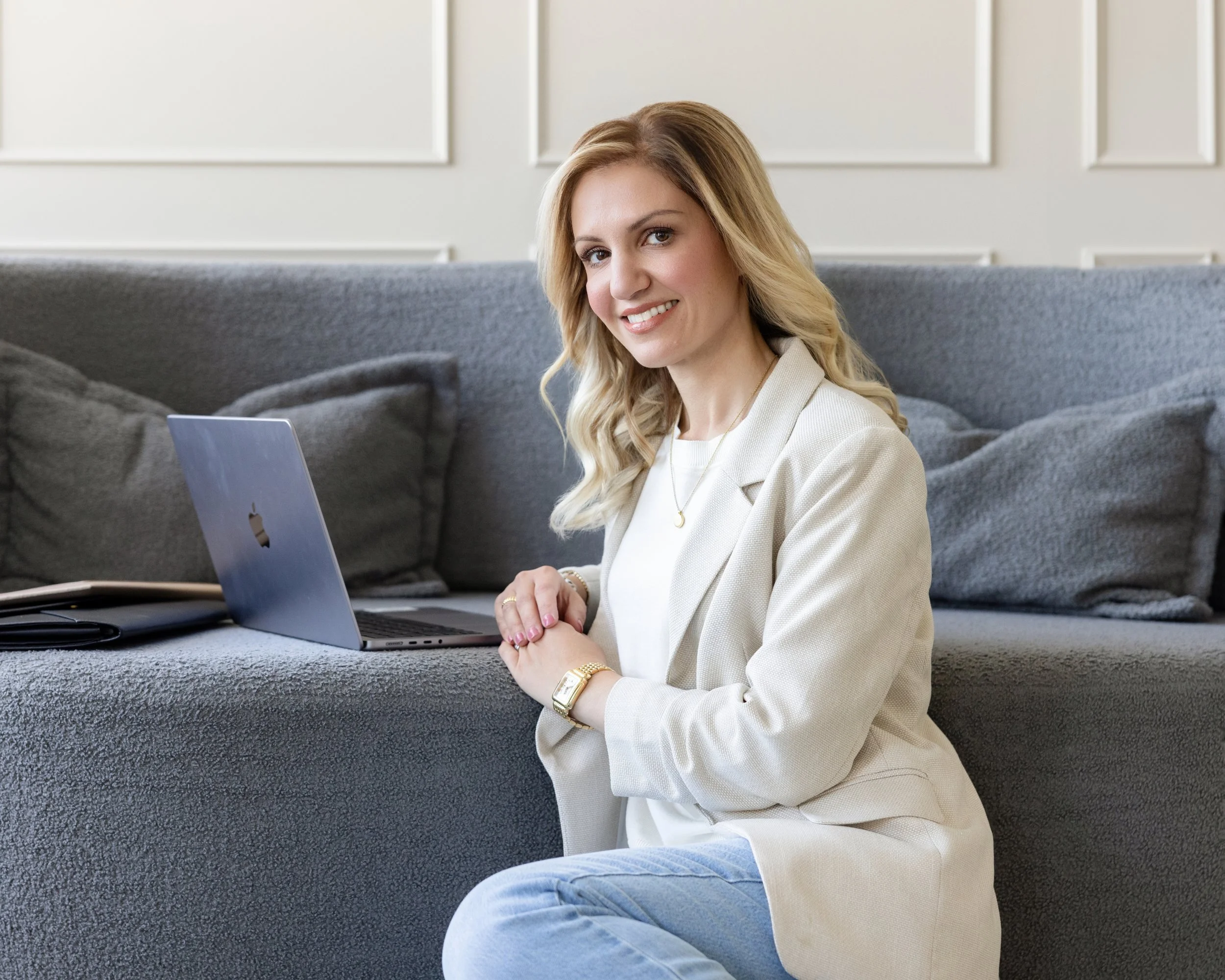 A woman with long blonde hair, wearing a beige blazer and white top, smiling while sitting on a gray couch with a laptop in front of her.