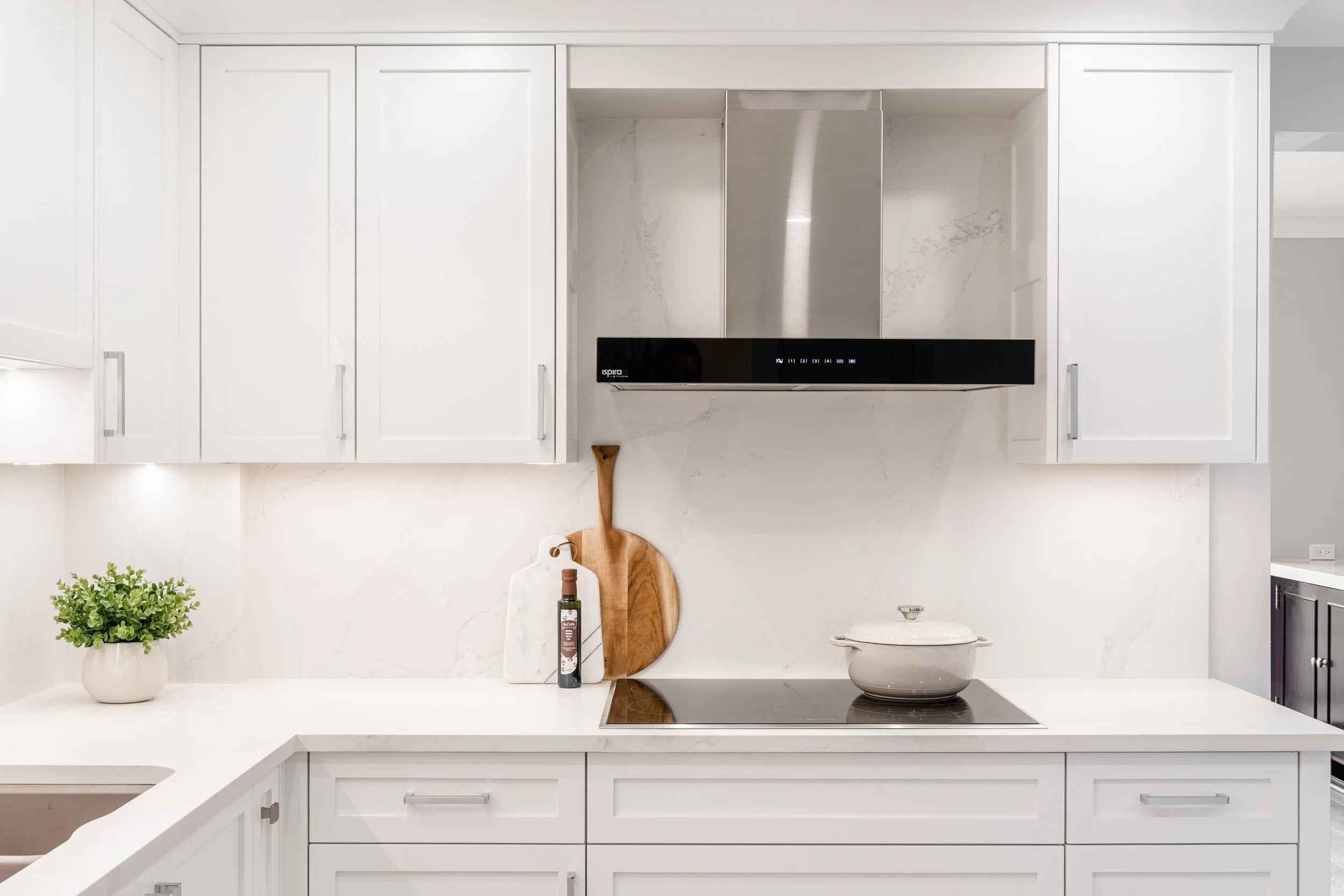 Modern kitchen with white cabinets, marble backsplash, induction cooktop, white ceramic pot on cooktop, cutting boards, plant in a pot, and stainless steel range hood.