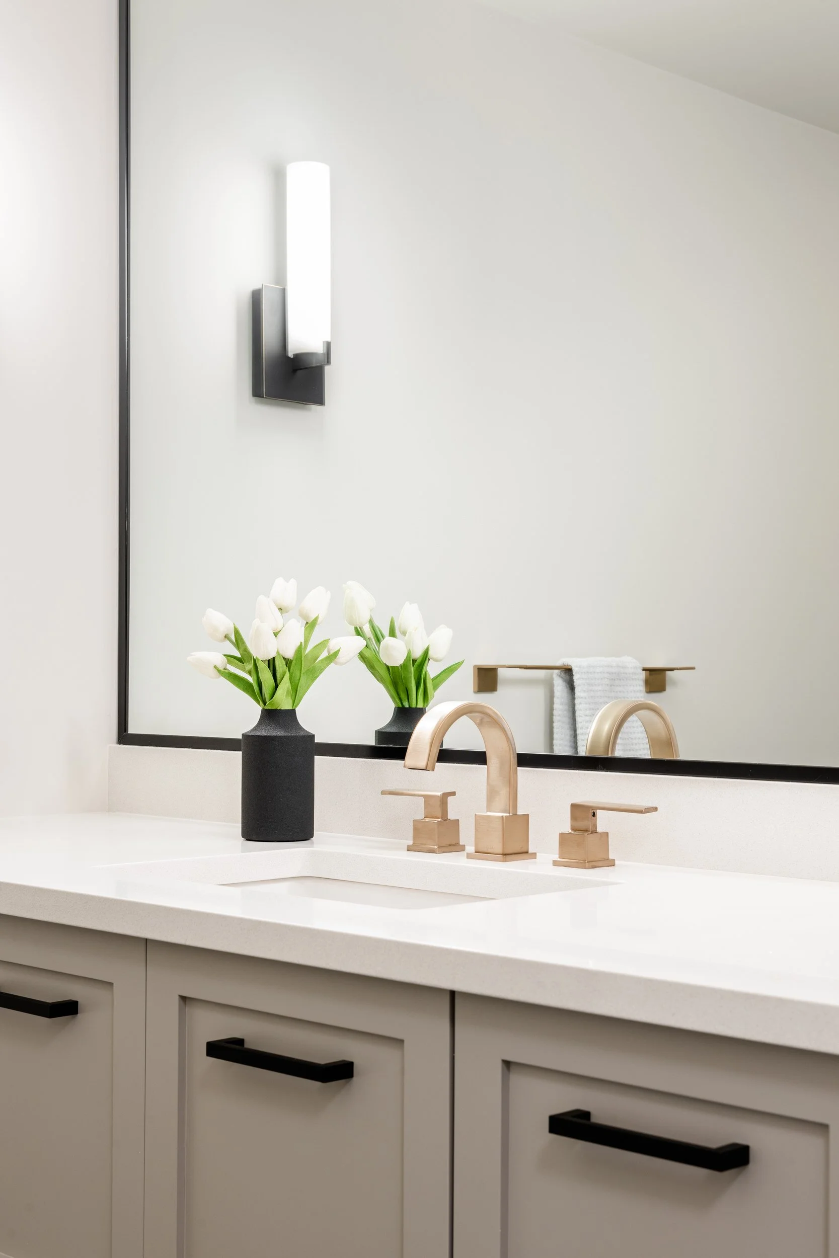 Bathroom vanity with white countertop, rose gold faucet, black vase with white tulips, large mirror, wall-mounted light, towel rack with white towel, and cream-colored cabinets with black handles.