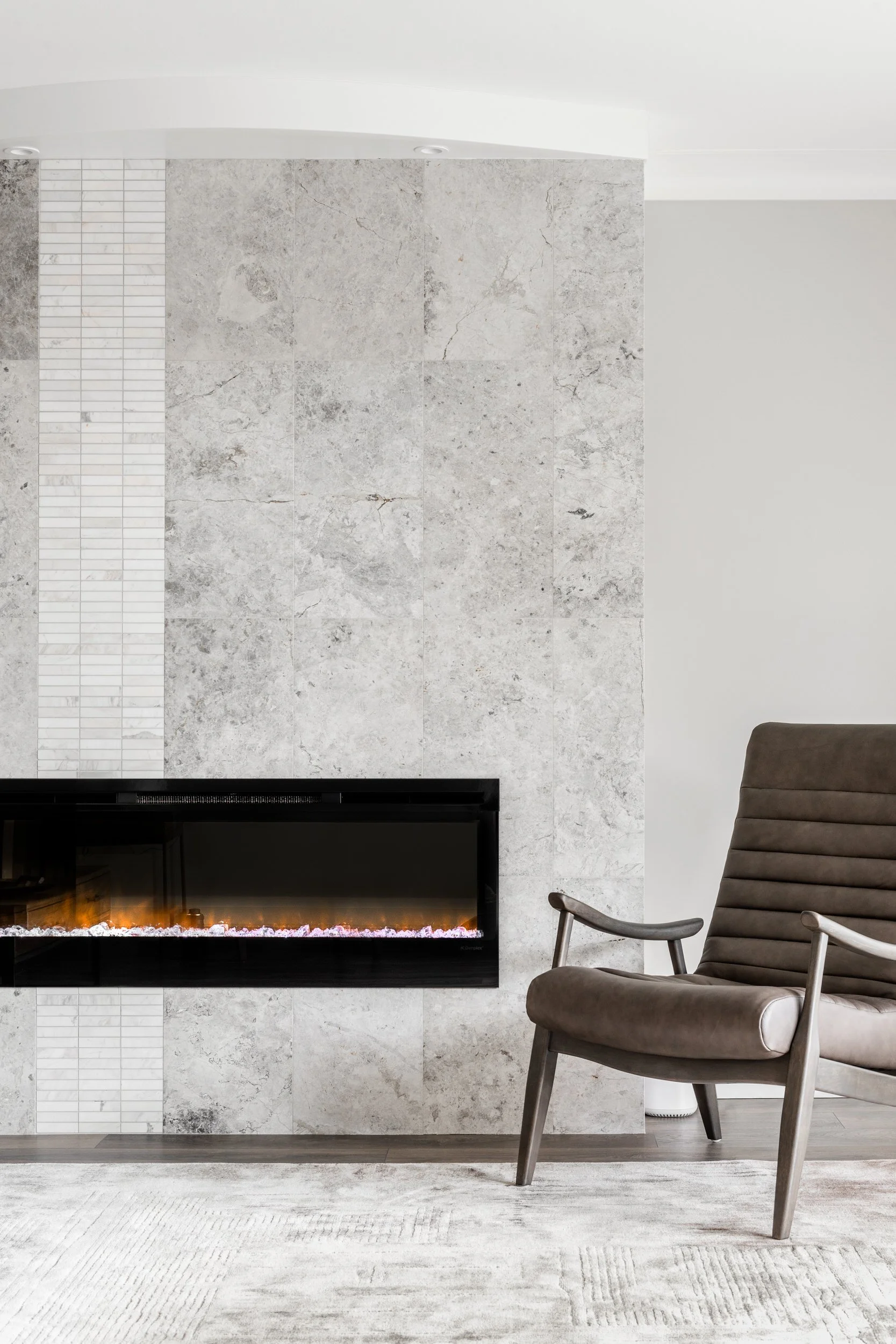 Modern living room with a grey stone fireplace, beige armchair with wooden armrests, and a white textured rug.