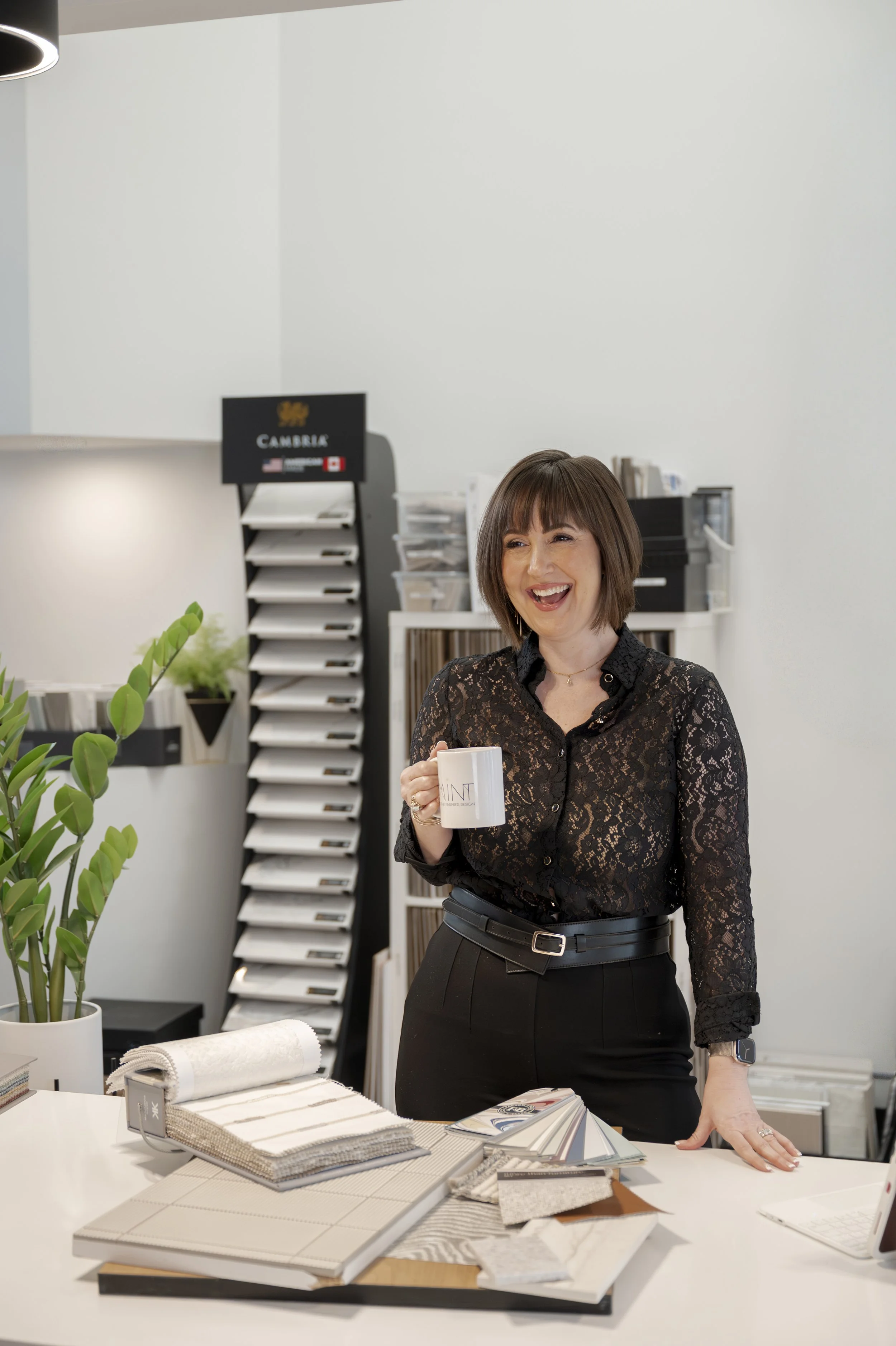 Principal Interior Designer Alexis Solomon, BID DID, smiling and holding a coffee mug in MINT Freshly Inspired Design studio in Victoria, BC, at a table with fabric and design samples.
