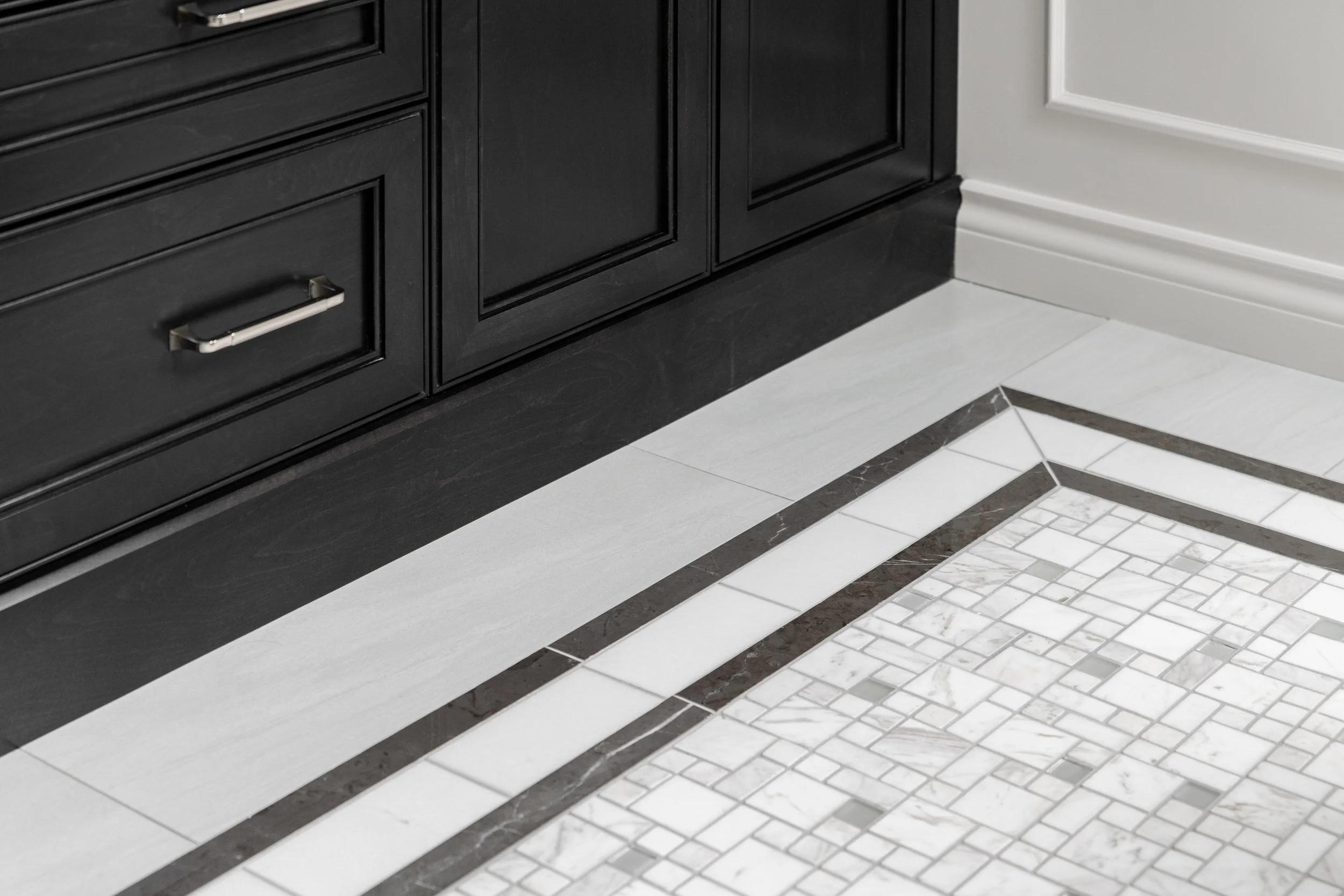 Close-up of a kitchen floor with a black cabinet, white and marble patterned tiles, and a decorative border of dark and light tiles.