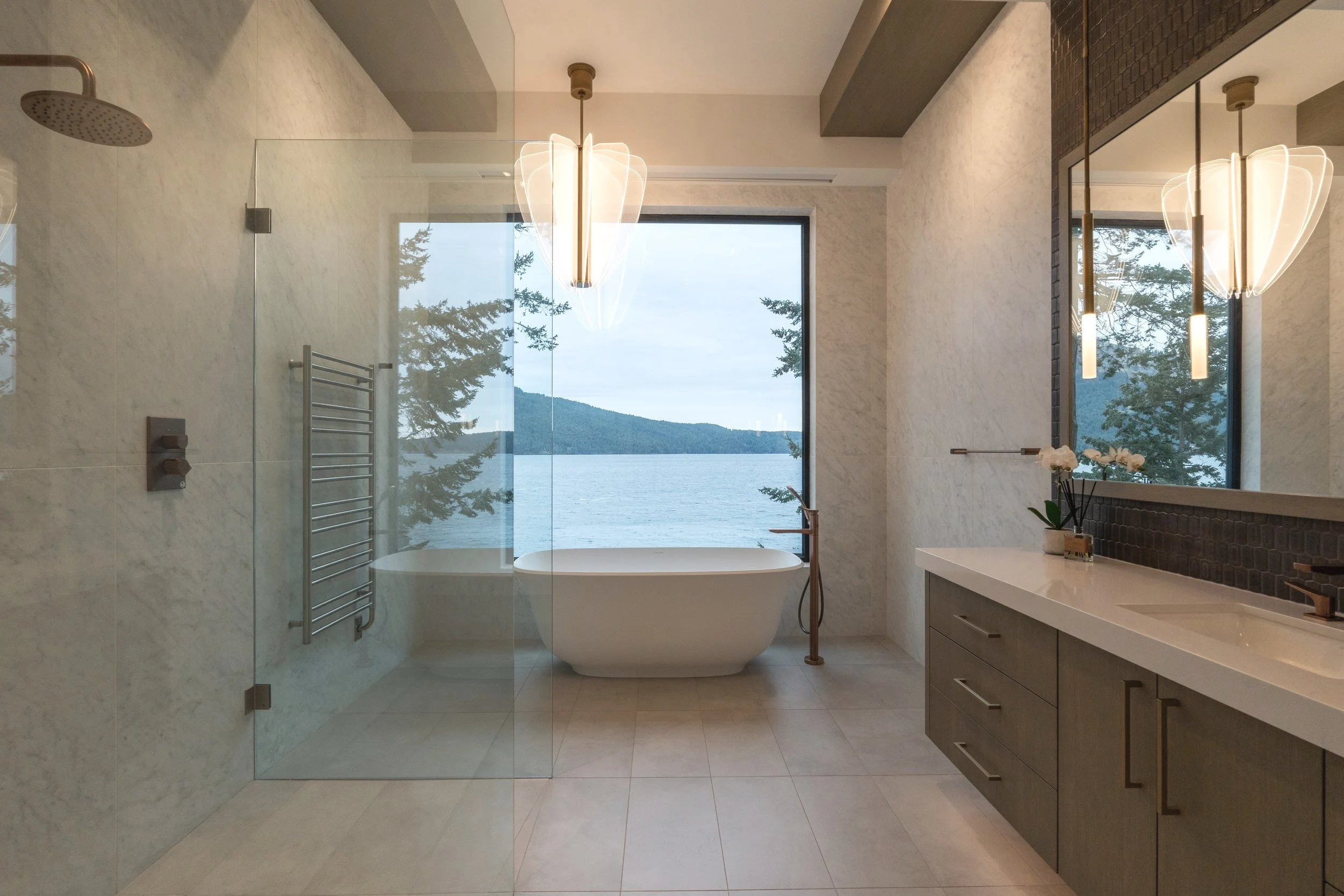 Modern bathroom with large window overlooking a lake, including a freestanding bathtub, glass-enclosed shower, and vanity with mirror and flowers.