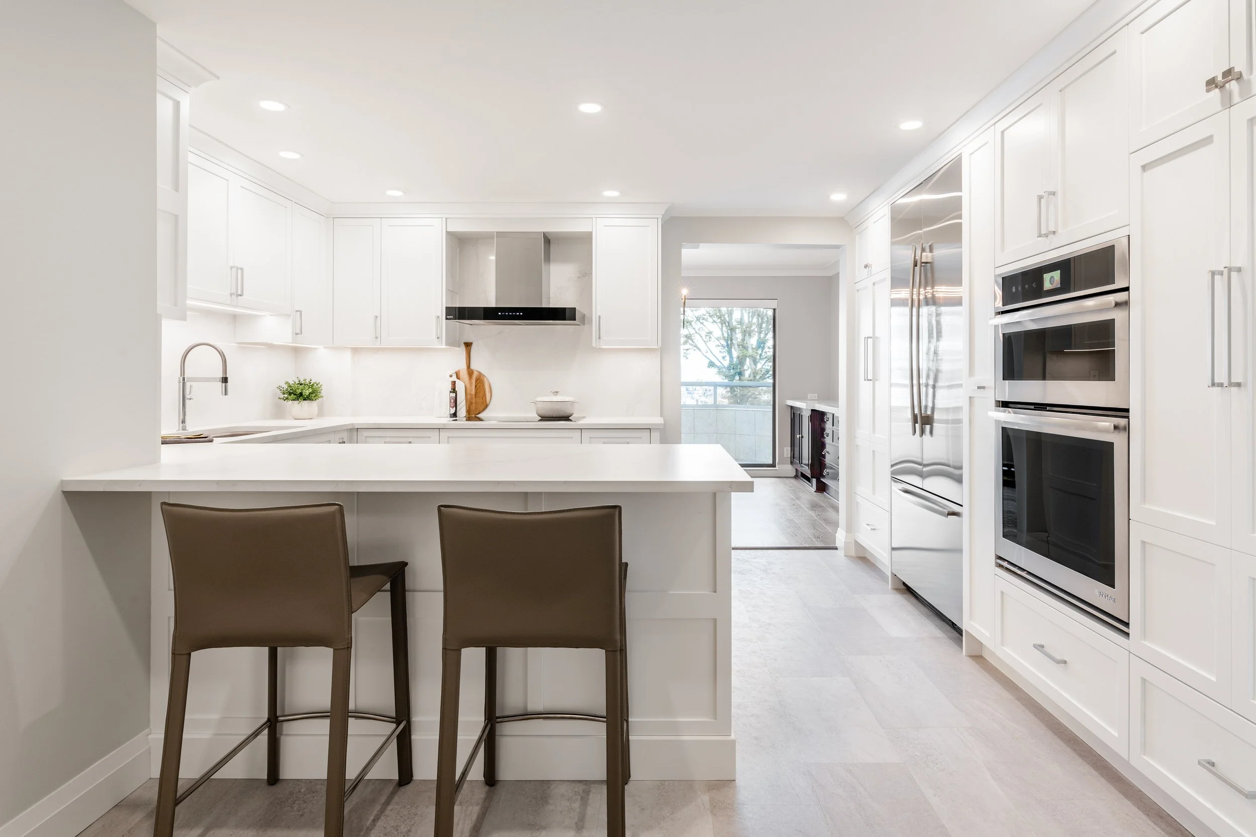 Modern white kitchen with island, brown bar stools, stainless steel appliances, and a sliding door leading to a balcony.