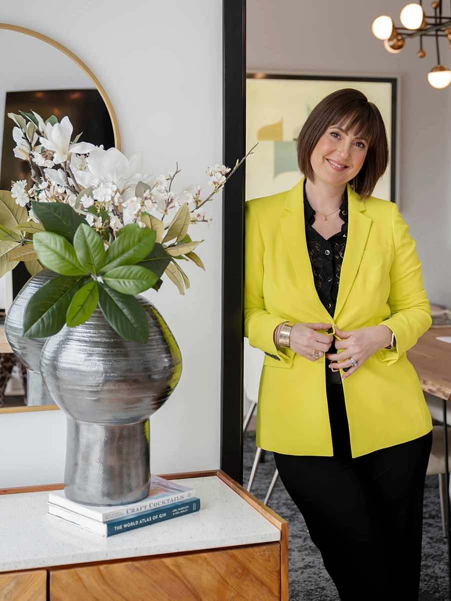Alexis Solomon, Principal Designer and Founder of MINT Freshly Inspired Design, a Victoria, BC interior designer, standing in a styled interior wearing a yellow blazer beside a decorative vase arrangement.