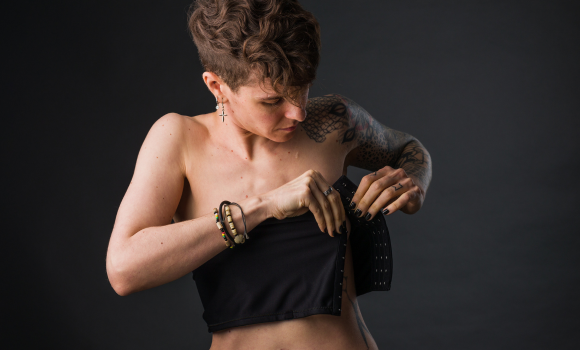 Trans person with short curly hair wearing bracelets adjusts their black binder against a dark background, symbolizing gender identity, support, and affirming care in therapy.