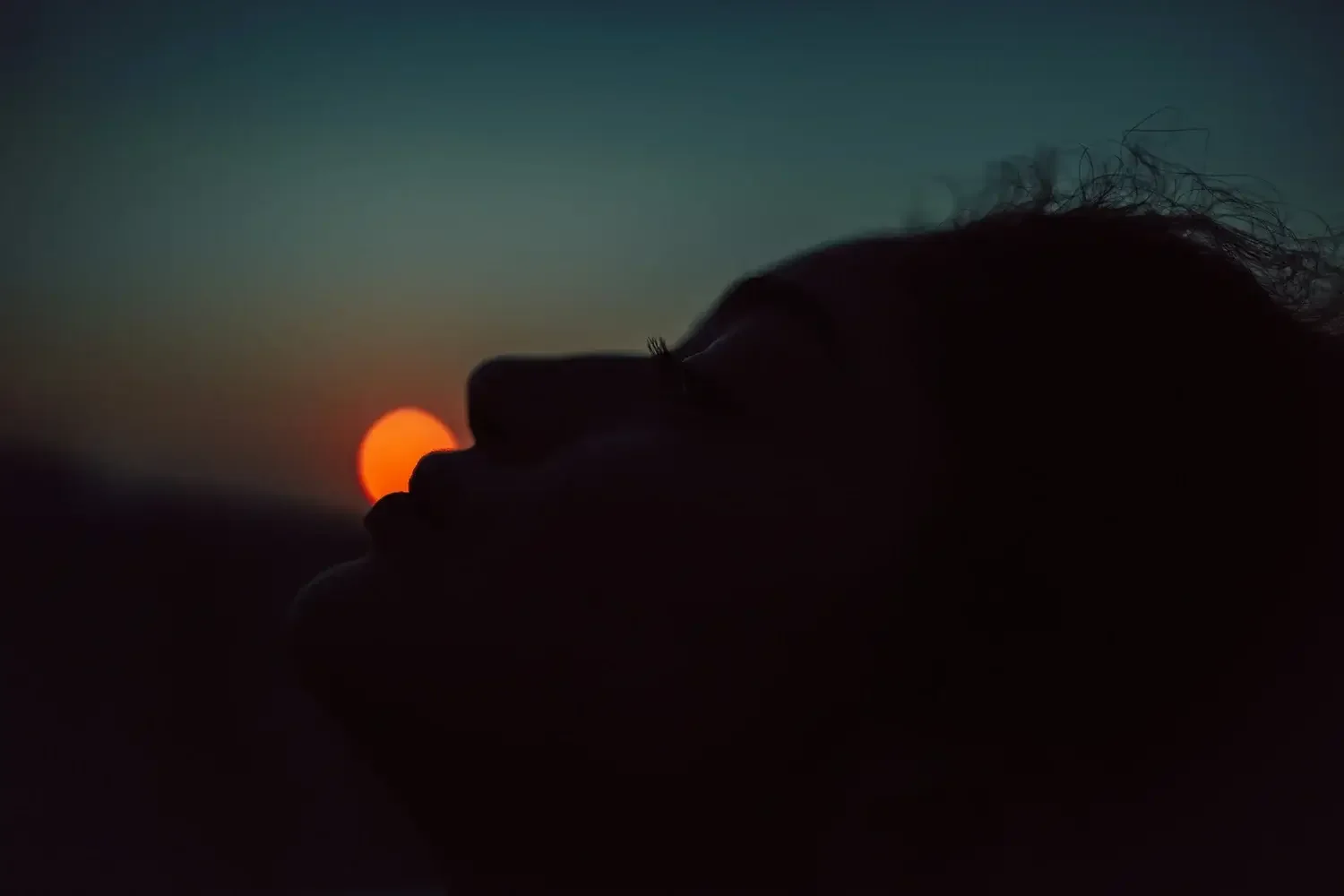 A person's silhouette with curly hair is lying down facing the sunset, with the sun near the horizon in the background.