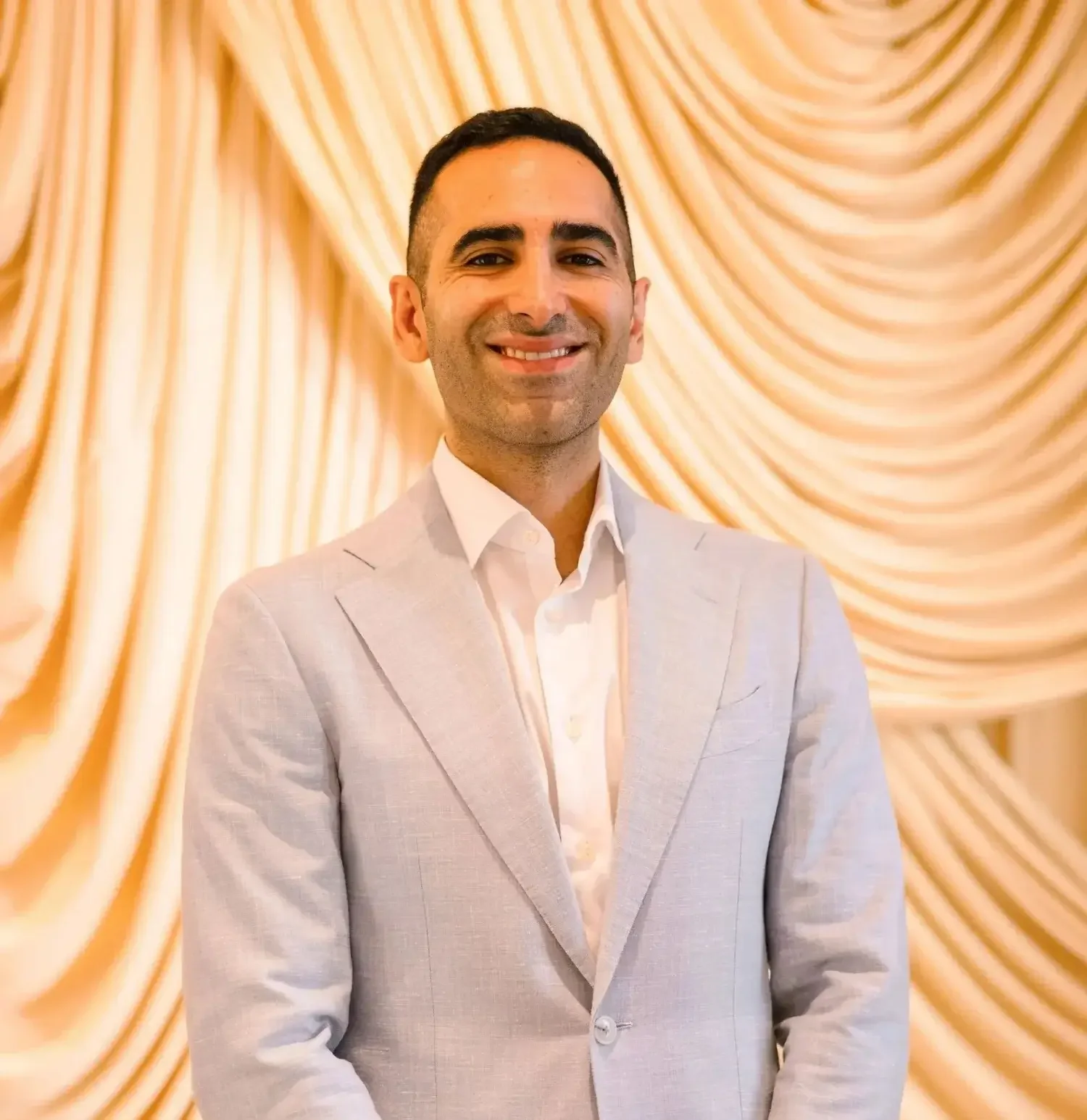 Dr. Nourian in a light blue suit is smiling in front of a golden curtain backdrop.