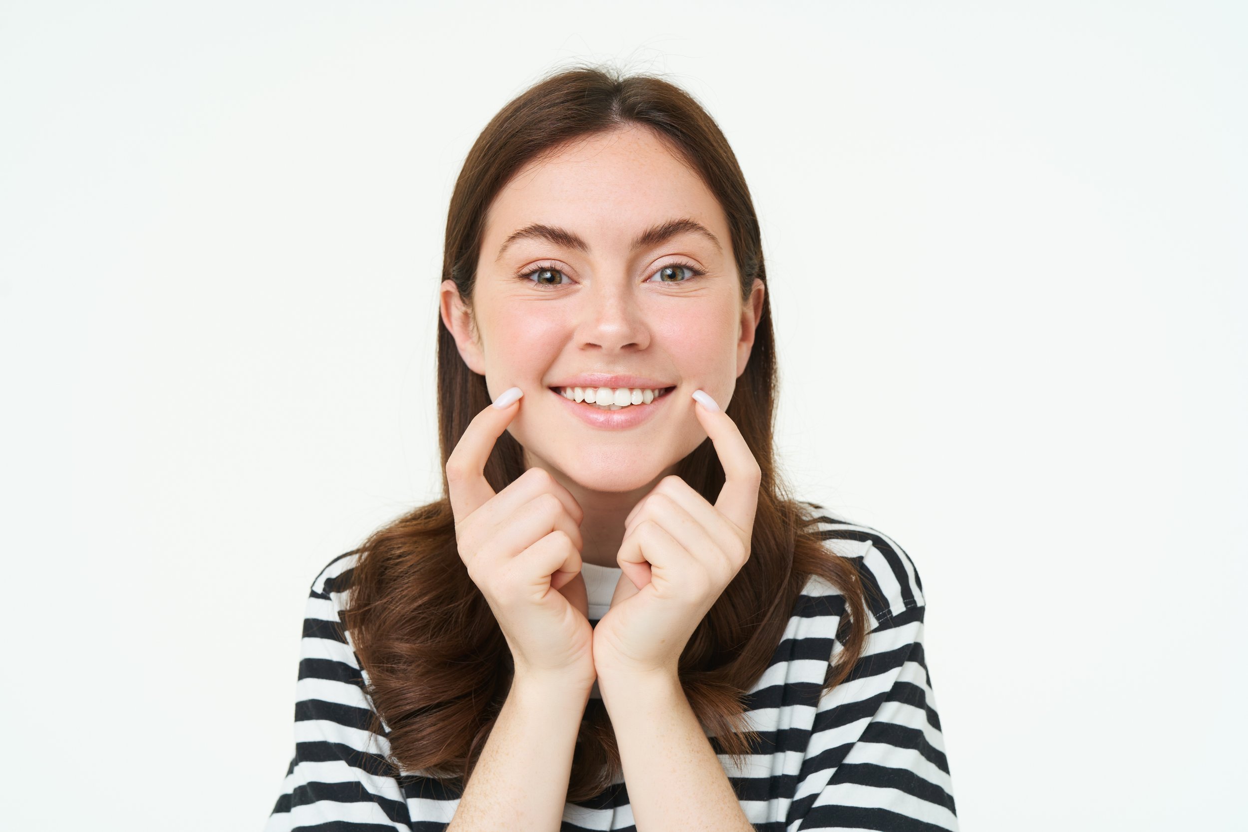 A young woman with long brown hair, wearing a black and white striped shirt, smiling and touching her cheeks with both hands.