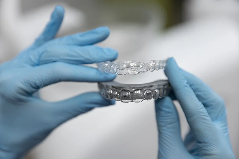 Close-up of hands in blue gloves holding clear dental aligners or retainers, showing two sets of transparent dental appliances.