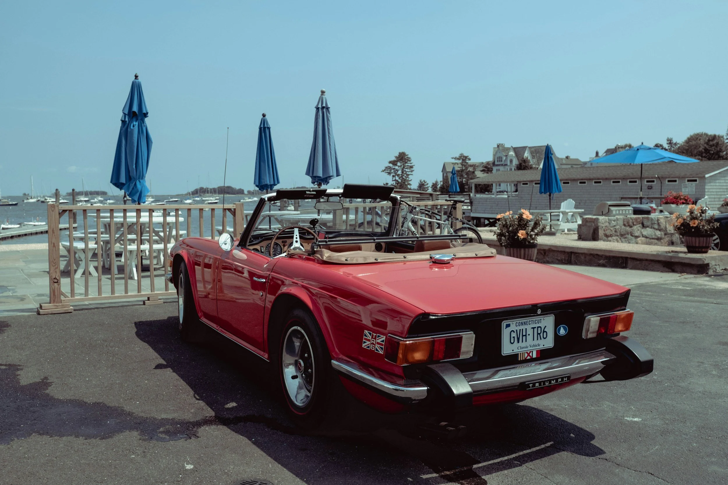 A red vintage convertible car with a Connecticut license plate parked near a marina with sailboats, blue umbrellas, and houses in the background.