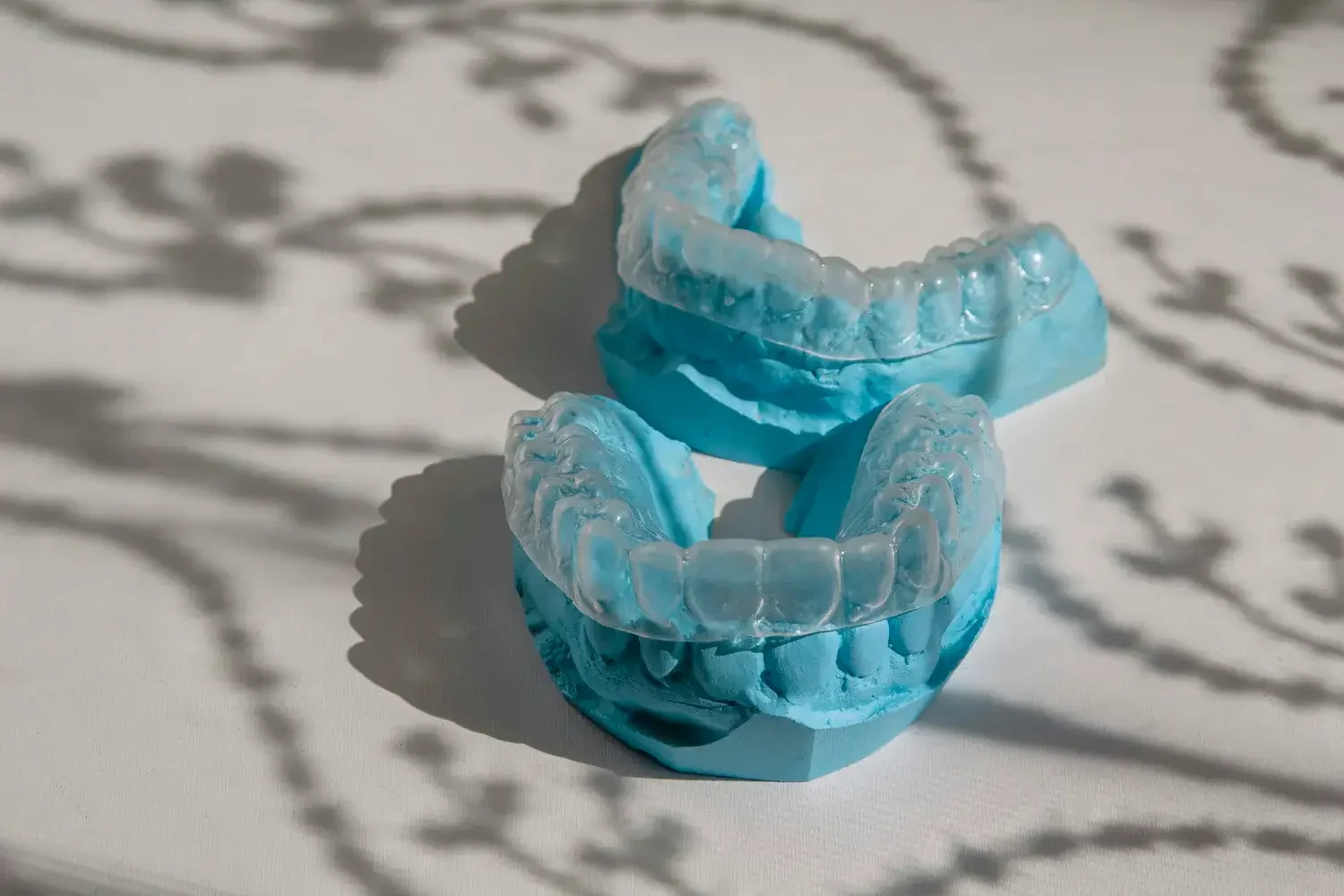 Two blue and clear dental retainers on a white surface with shadows of decorative plant shadows.