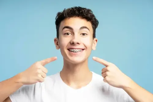 Young man smiling and pointing at his teeth with both index fingers against a light blue background.
