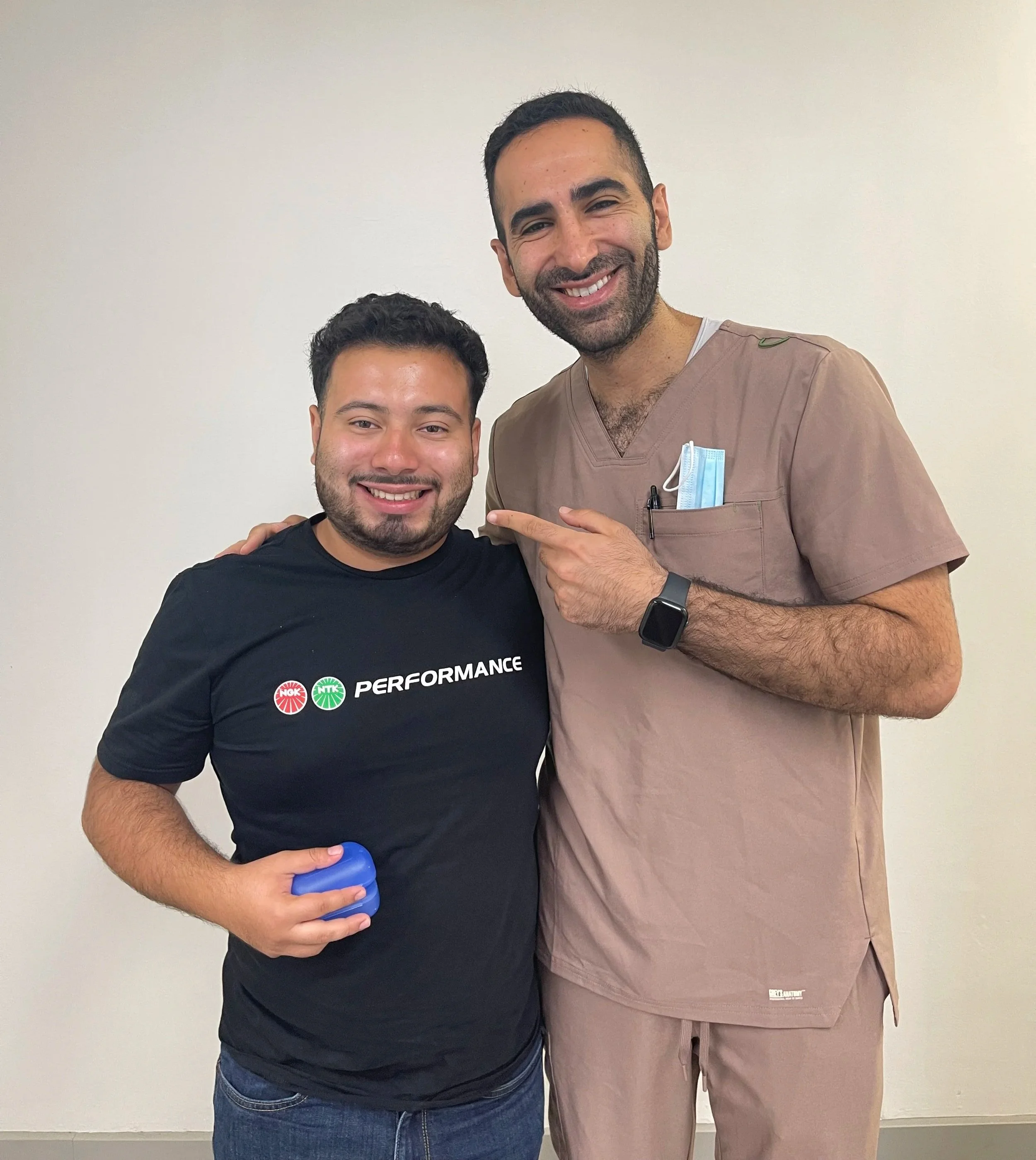 Two men smiling and posing together, one wearing a black T-shirt with the word 'PERFORMANCE' and two logos, the other in medical scrubs with a mask in the pocket, against a plain white wall.