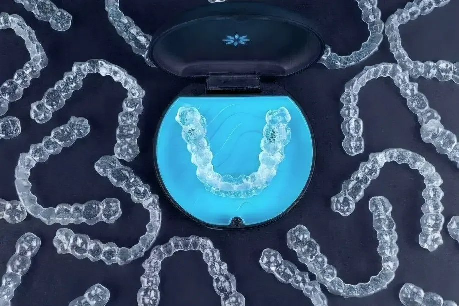 An open dental retainer case with a clear aligner inside, surrounded by transparent, flexible plastic aligners spread out on a dark surface.