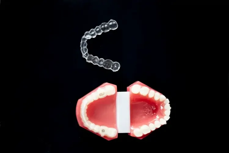 A set of artificial teeth with gums, a white strip representing dental floss, and a clear plastic dental brace or retainer above them, displayed against a black background.