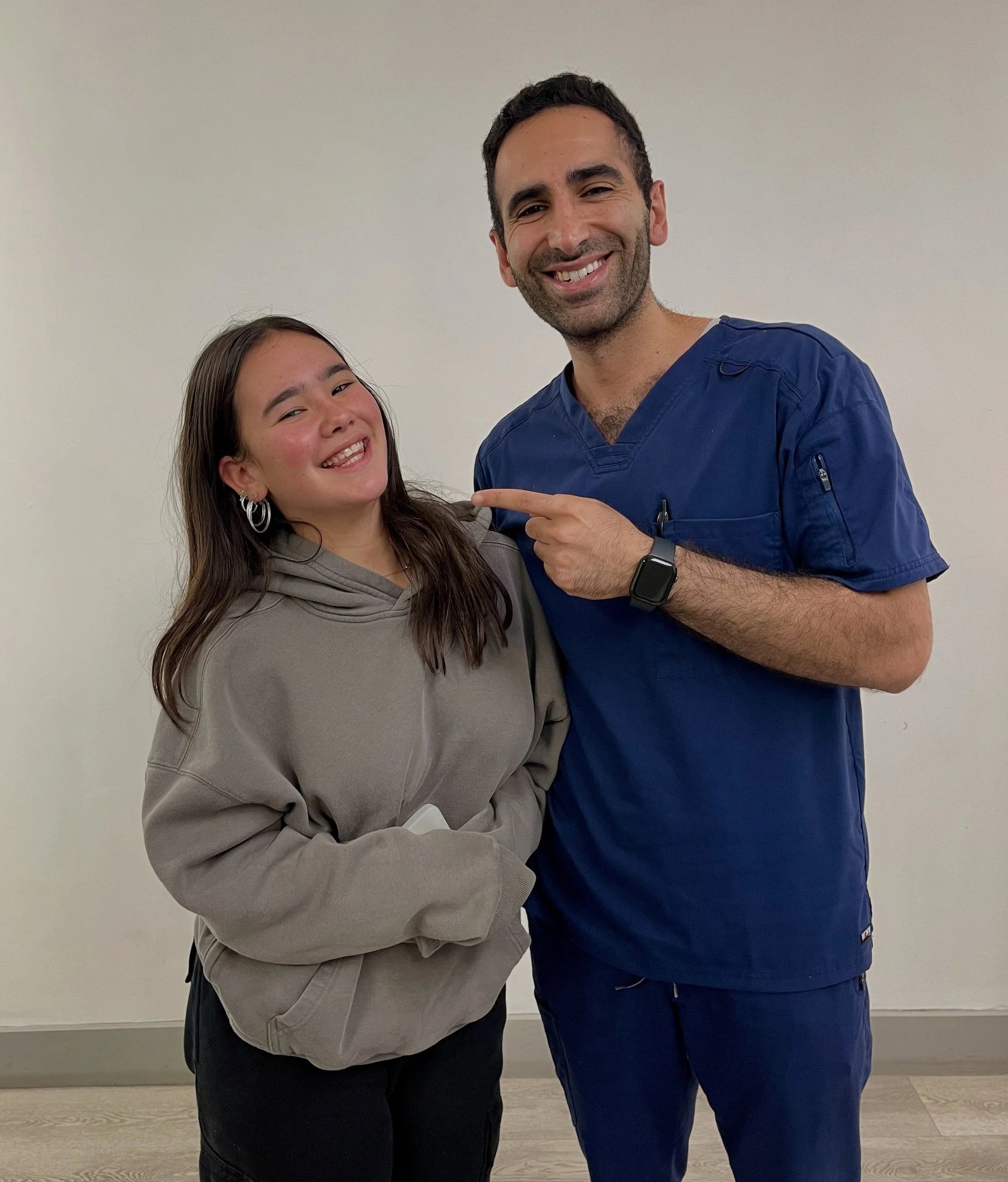 A woman and a man in medical scrubs smiling and posing together. The man is pointing at the woman.