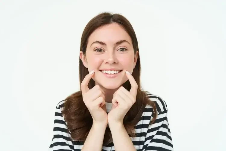 A young woman with brown hair smiling and pressing her fingers to her cheeks, wearing a black and white striped shirt against a white background.