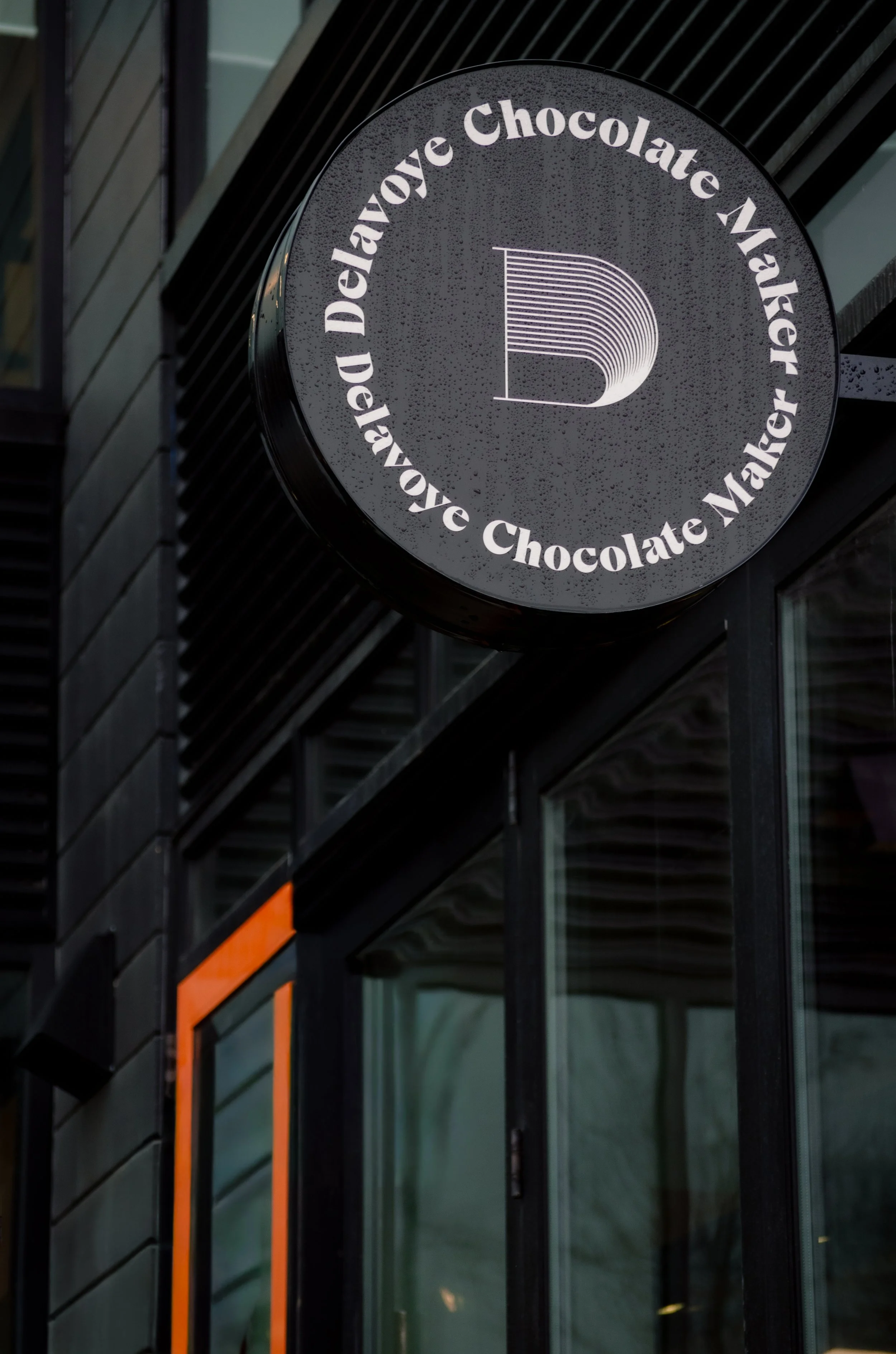 Round black sign with white text that reads 'Delavoye Chocolate Maker' and a stylized letter 'D' logo in the center, mounted on a black wall with diagonal lines and windows visible in the background.