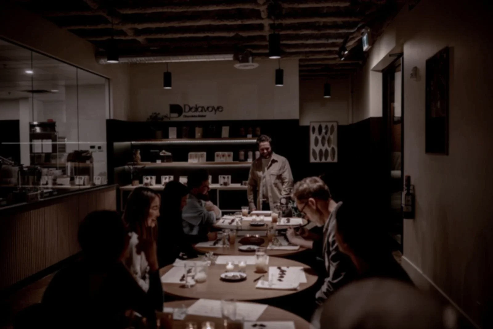 People dining at tables in a dimly lit restaurant or cafe with wooden ceiling beams and modern lighting fixtures.