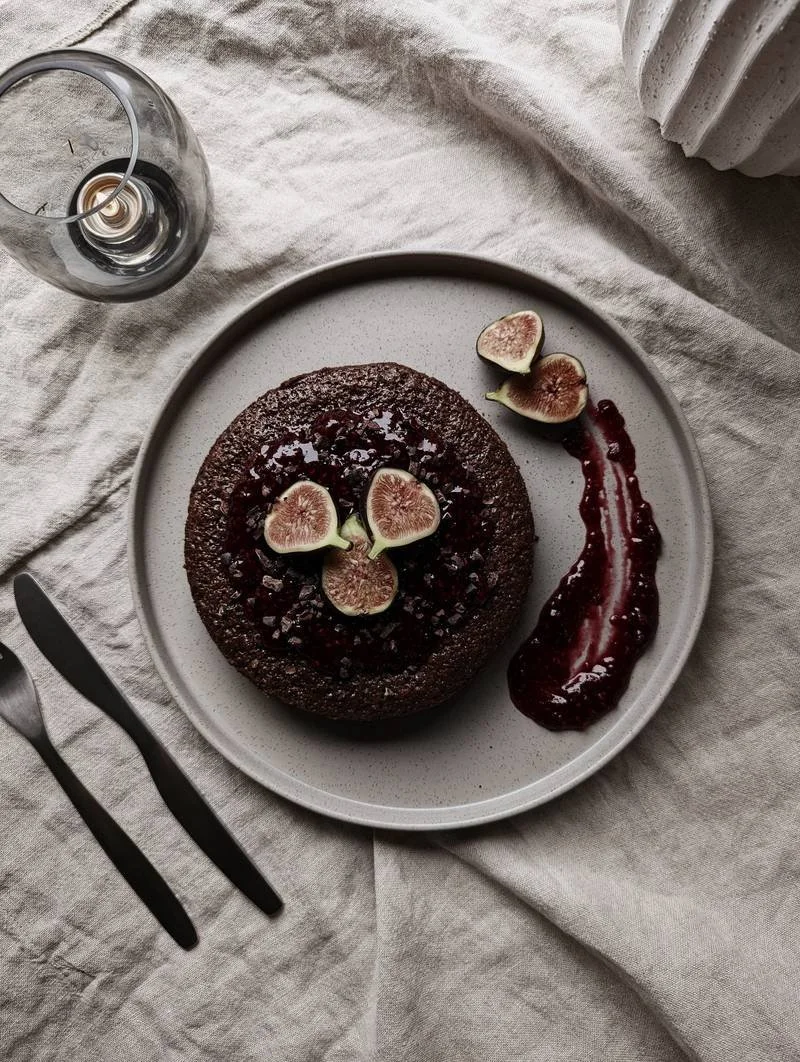 A chocolate cake topped with fig slices and a berry sauce on a white plate, with a glass of water and a fork on a textured tablecloth.