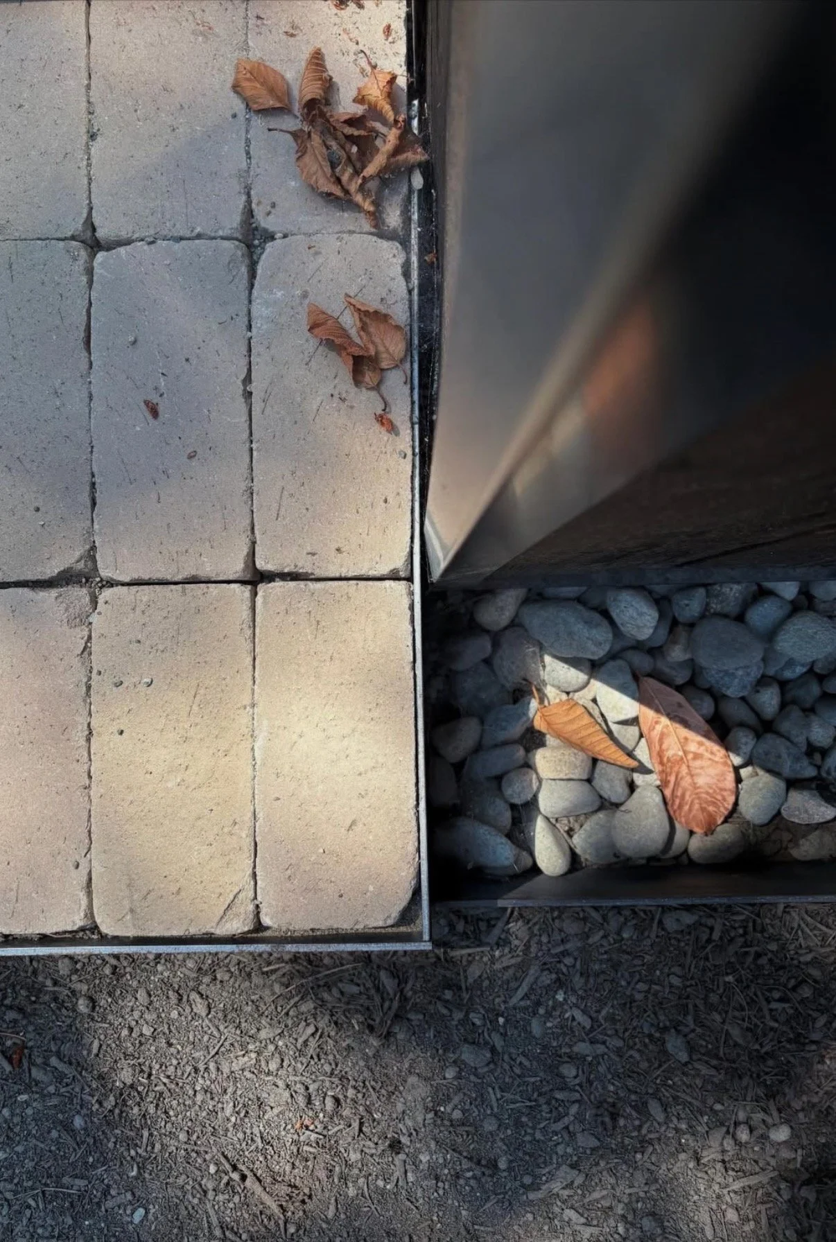 Close-up of an exterior material transition between concrete pavers, metal edging, drainage rock, and a vertical architectural element on a custom residential project.