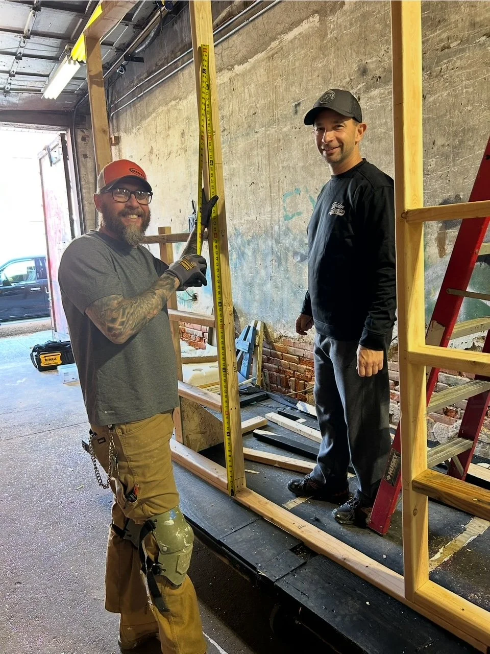 Two men working on a construction project inside a garage or workshop. One man is holding a measuring tape vertically, smiling and wearing a red cap, glasses, and work pants. The other man is standing on a wooden platform, smiling and wearing a black cap and black sweatshirt. There are wooden frames, a red ladder, and tools around them.