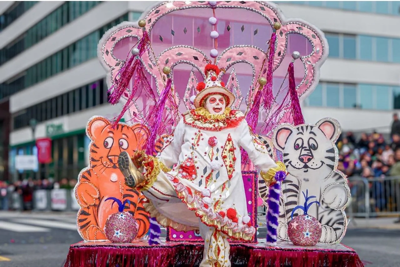 Person dressed as a clown with face makeup, wearing a red, white, and gold costume, sitting on a decorative float with cartoon tiger and zebra cutouts, during a parade on a city street with buildings and spectators.