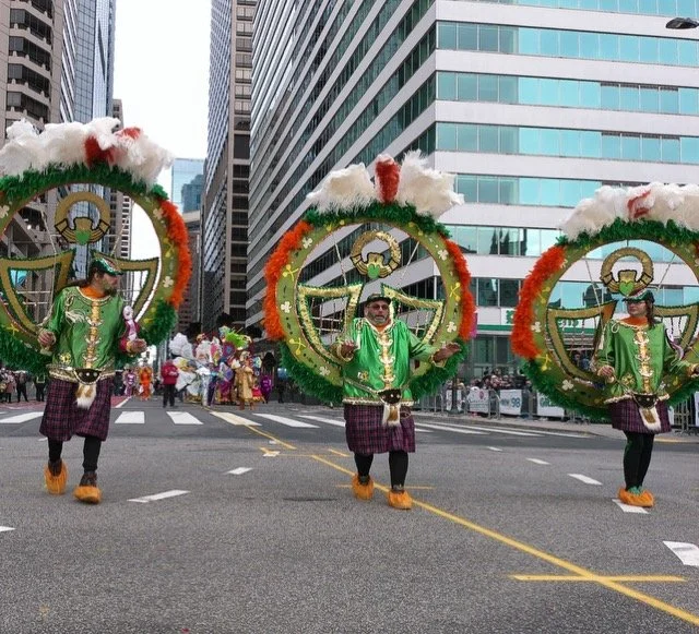 Parade with three performers in green costumes and large circular feathered headdresses decorated with Christmas ornaments, strutting on city street with tall modern buildings in background.