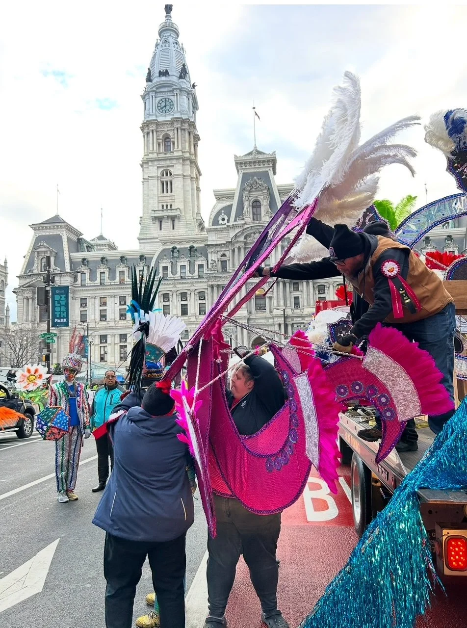 People working on a float decorated with colorful feathers and sequins during a parade in front of a historic building with a clock tower.