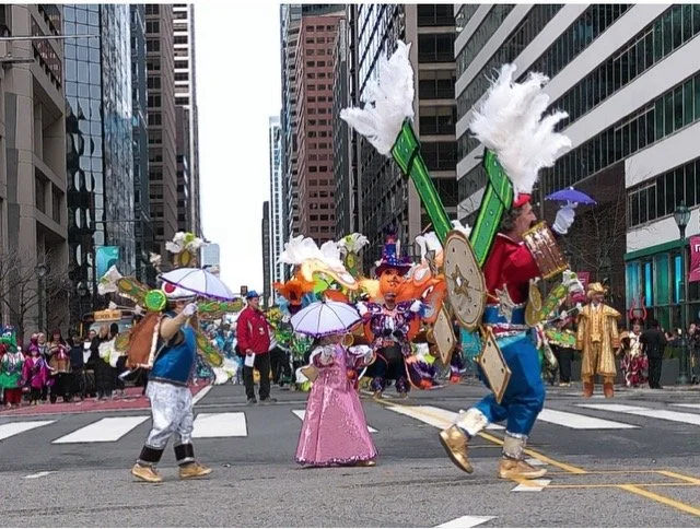 People in colorful costumes and hats participating in a parade on a city street, carrying umbrellas and large decorative props.