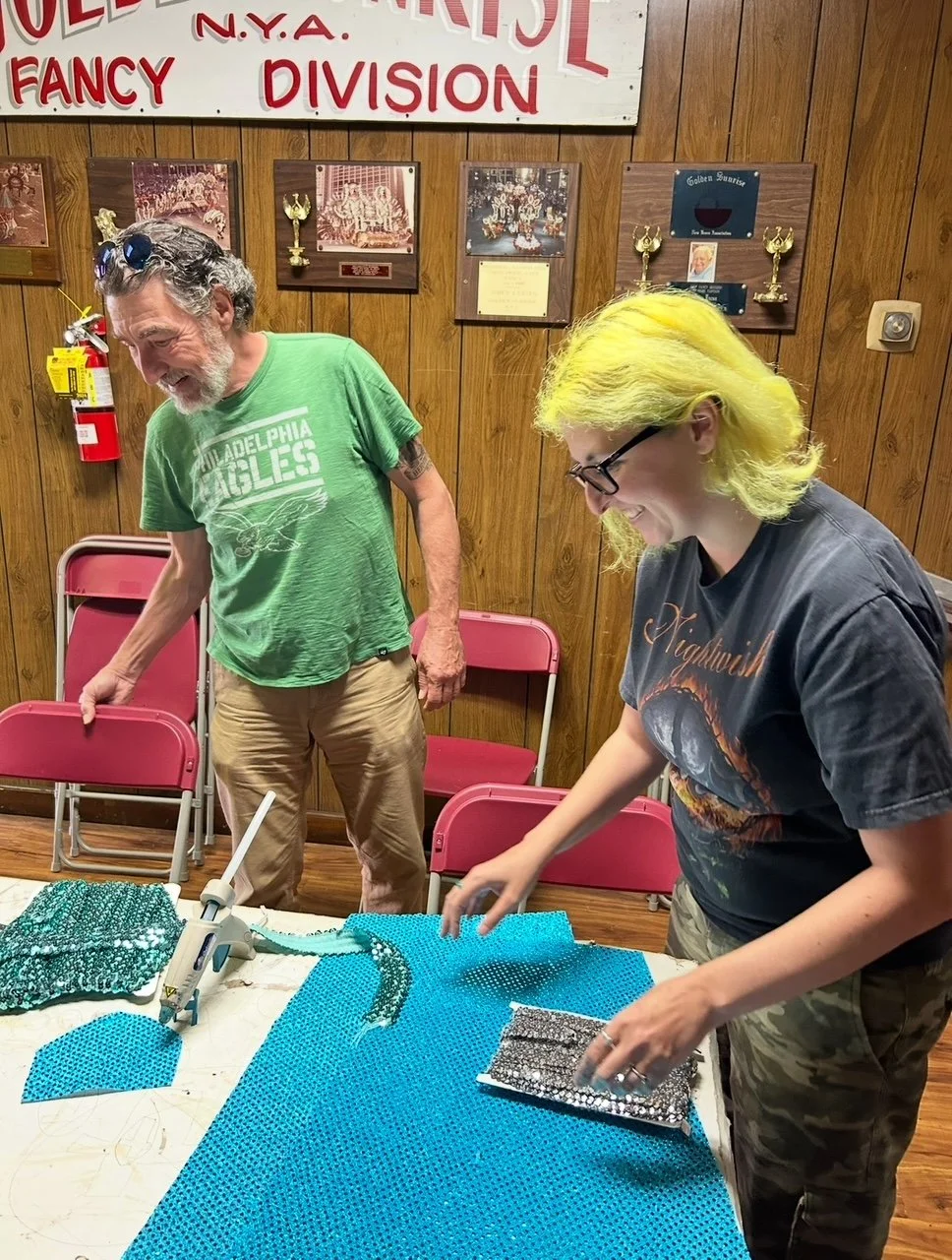 Two people working on a mosaic project at a table inside a room with wooden walls. The person on the right has yellow hair, wears glasses, and a dark T-shirt. The person on the left has gray hair, a beard, and wears a green Philadelphia Eagles T-shirt. There are pink chairs stacked against the wall and framed trophies and photographs on the wall, with a sign above that reads "N.Y.A. Fancy Division."