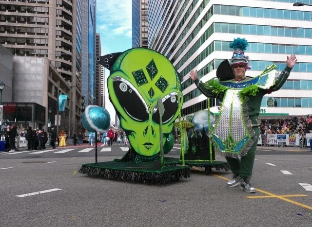 Float in a parade featuring a large green alien head with black eyes and a small black spaceship on its forehead, accompanied by a person in a colorful, shiny, elaborate costume.