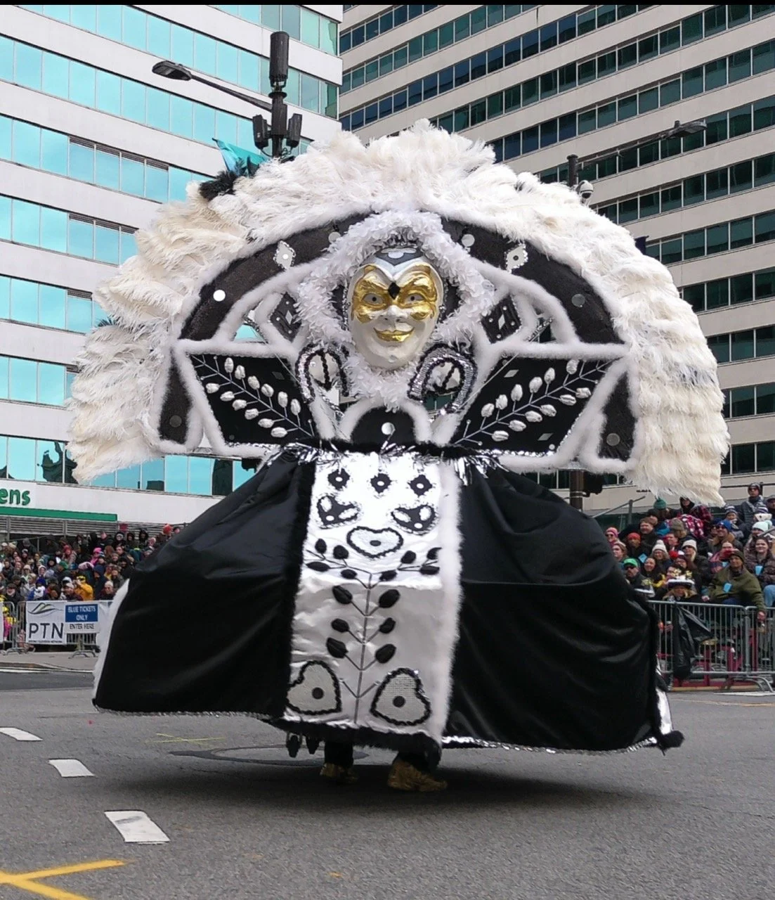 A person in a large, elaborate costume with a smiling face mask, decorative feathers, black and white fabric, and intricate patterns, participating in a parade in an urban area with tall buildings and a crowd of spectators.