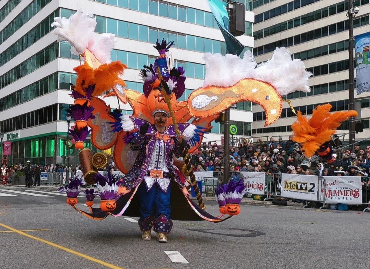 A carnival parade with a performer in colorful, elaborate costume in front of a crowd and city buildings.