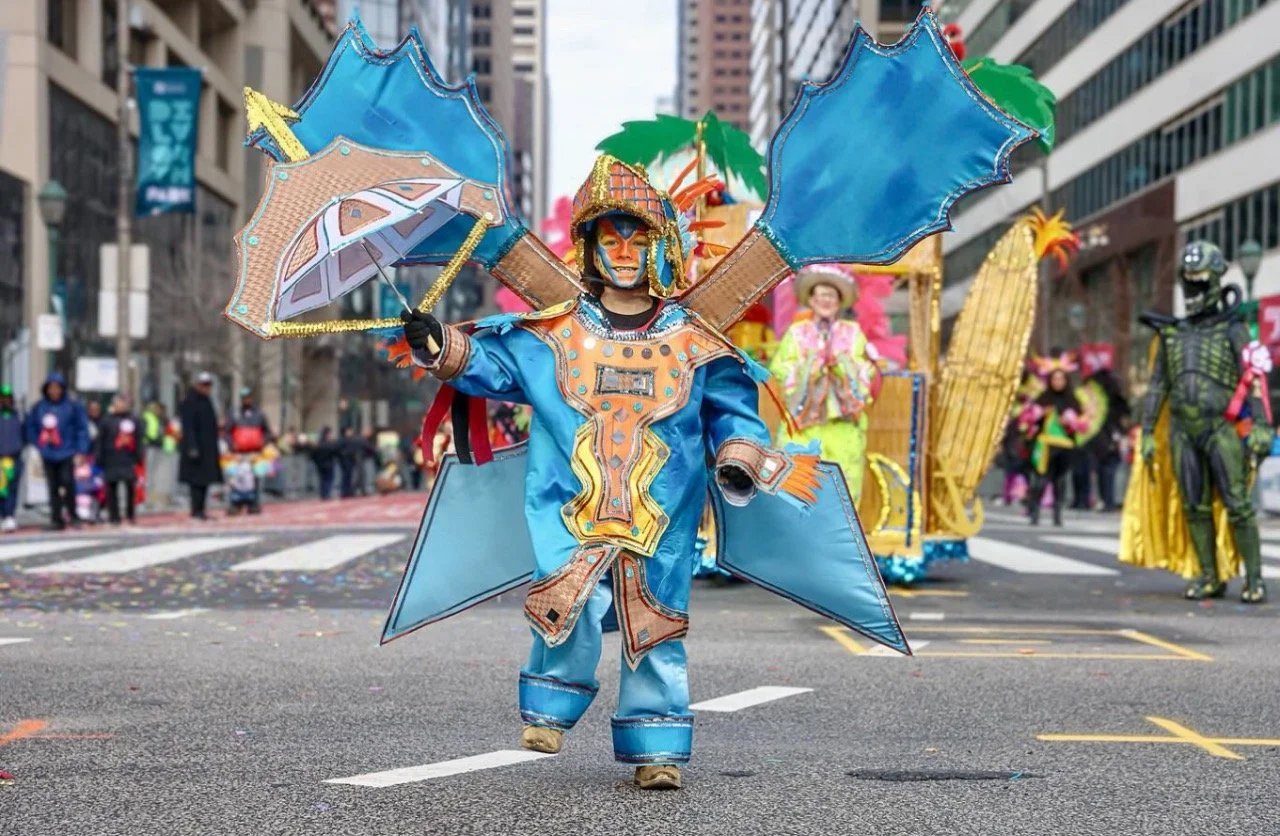 A person dressed in a colorful, elaborate costume with large wing-like structures and holding umbrellas, participating in a parade on a city street.
