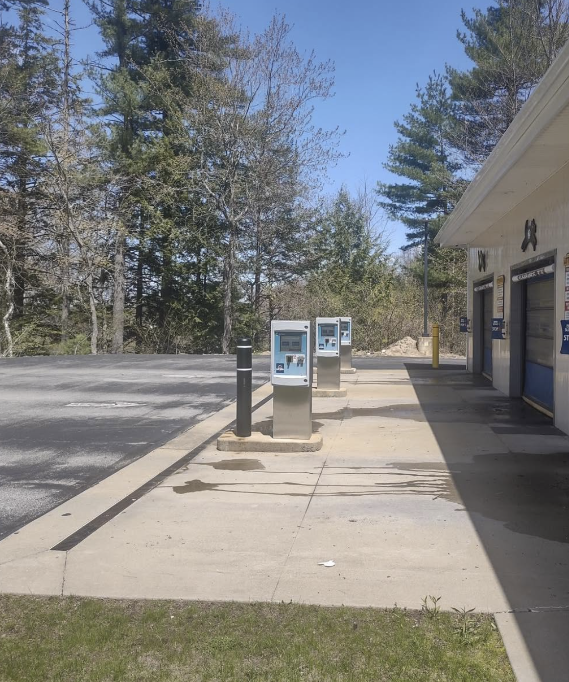 An outdoor parking lot with three electric vehicle charging stations next to a building, backed by trees and a clear blue sky.