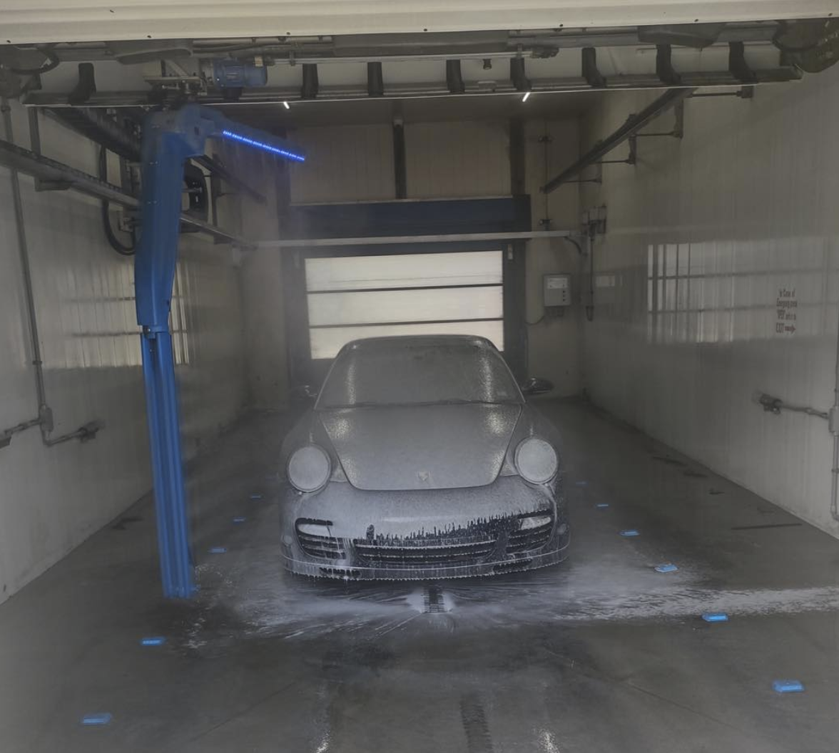 Car in an automated car wash with soap foam and water spray, with a blue robotic arm washing the vehicle.