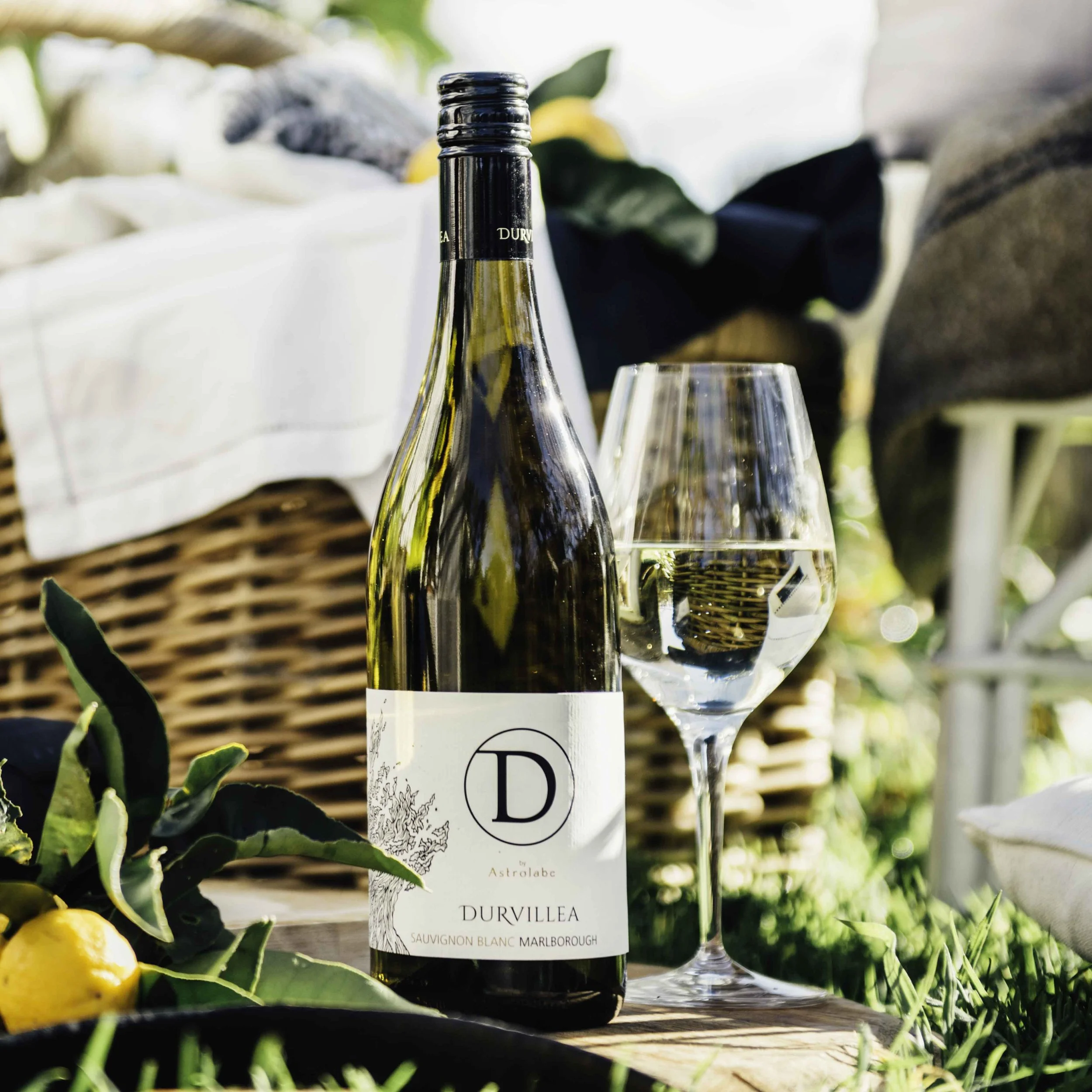 Durvillea Sauvignon Blanc bottle and wine glass outdoors on a picnic setting with lemons, wicker basket, and summer greenery.