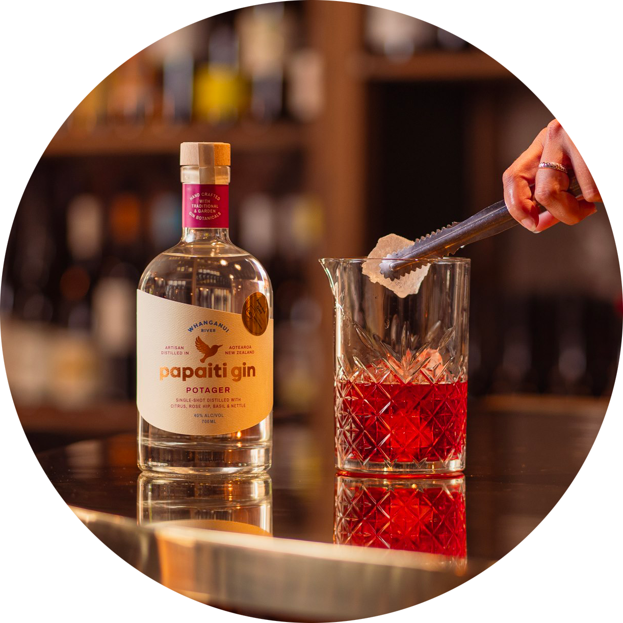 Papaiti Potager Gin bottle beside a crystal glass with red cocktail as ice is placed into the drink in a warm bar setting