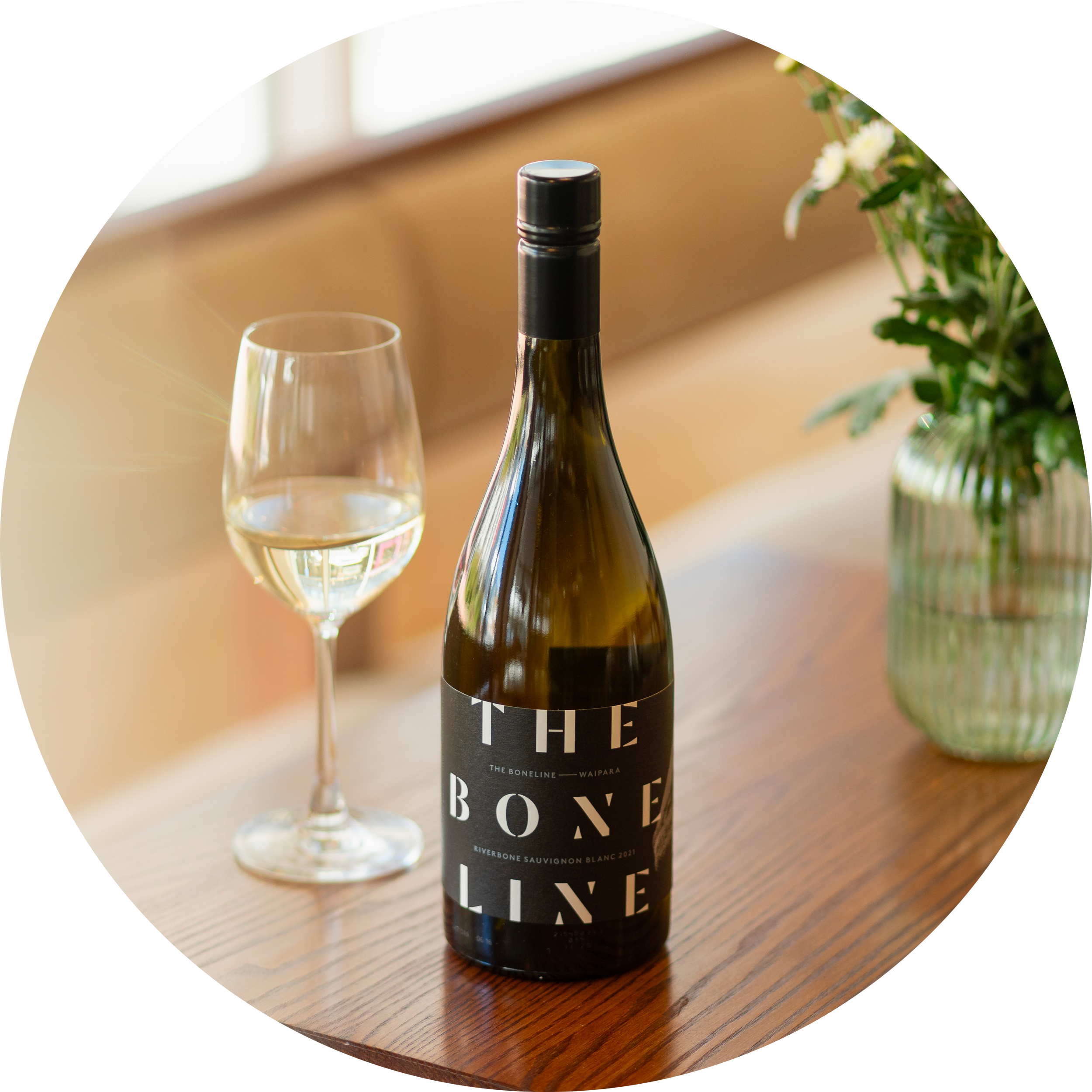 Bottle of The Bone Line Sauvignon Blanc on a wooden table beside a filled wine glass in natural window light