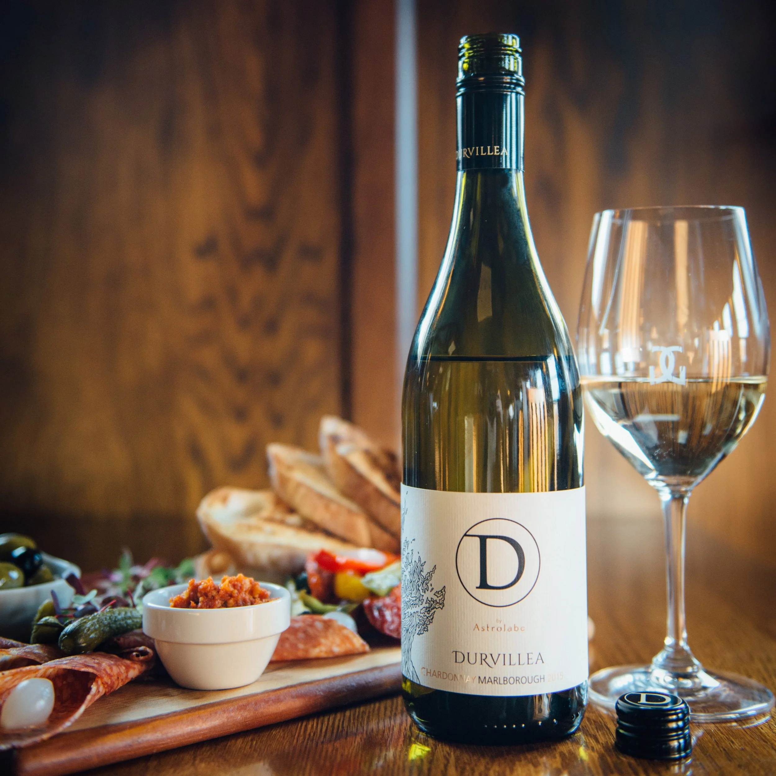 Durvillea Chardonnay bottle with a glass of wine beside a charcuterie board featuring olives, breads, and cured meats.